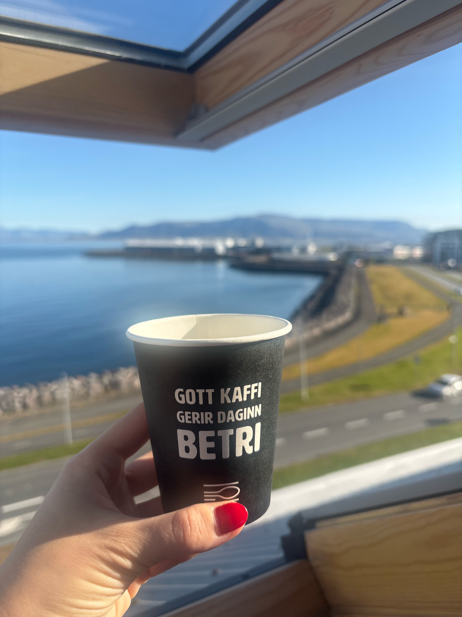 A hand with red nail polish holds a black coffee cup with white text that reads "GOTT KAFFI GERIR DAGINN BETRI" which translates to "Good coffee makes the day better." In the background, the ocean meets the coastline of Reykjavik, Iceland, with mountains visible in the distance.