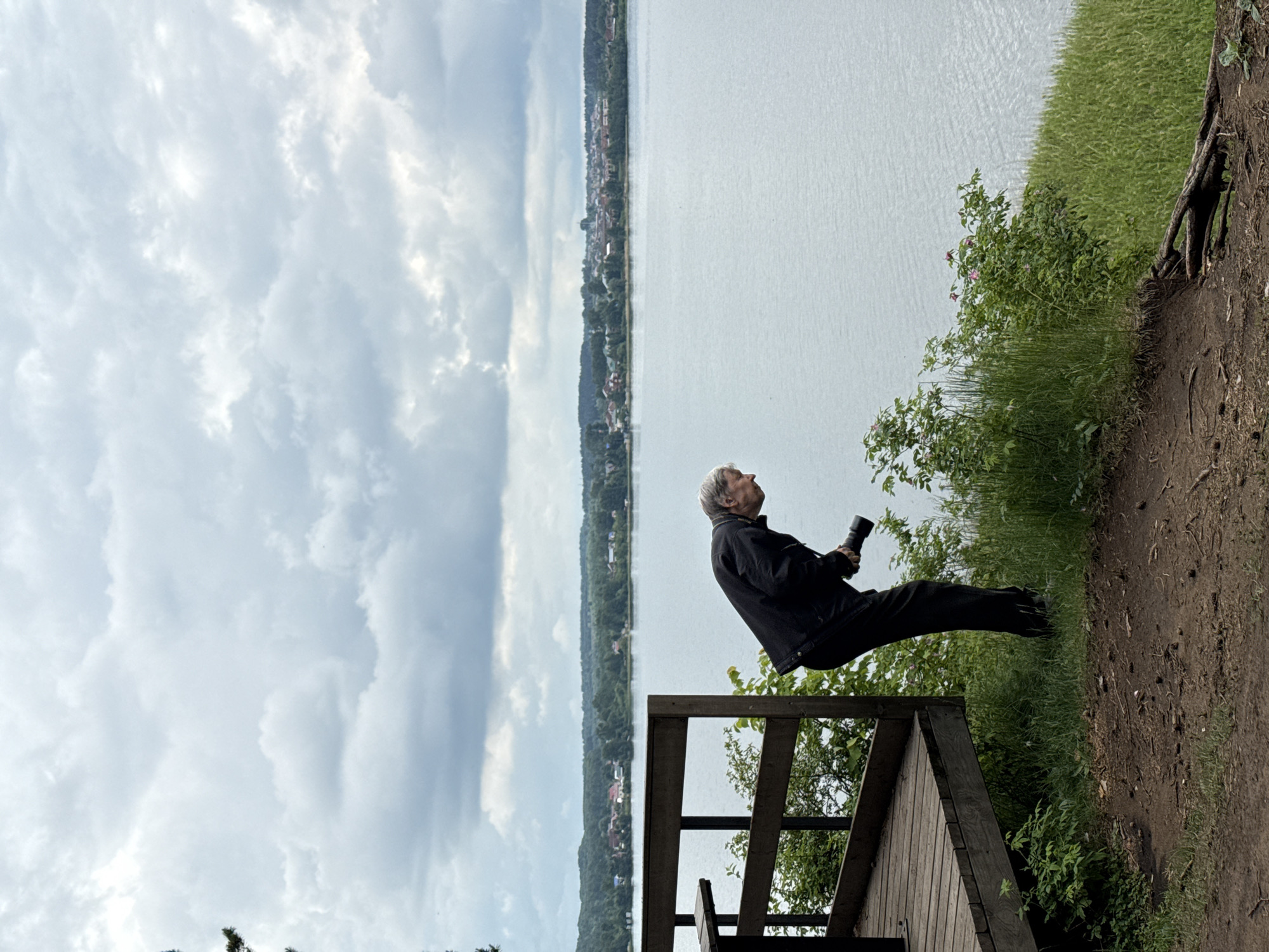 An outdoor, horizontally oriented image depicts an older man observing a distant scene over a large body of water on an overcast day in Votkinsk, Russia. The man, with gray hair, is dressed in a dark jacket and dark trousers. He stands on a wooden observation deck or platform, looking upwards and to his right (viewer's left in the correctly oriented image), holding a black monocular or a small camera with a telephoto lens in his right hand, suggesting focused observation.
In the midground, a wide body of water, consistent with the Votkinsk Reservoir, stretches across the frame, its surface displaying subtle ripples. The distant shoreline across the water is densely covered with green trees, behind which a settlement with houses or buildings is visible. The sky above is filled with light gray and white clouds, indicating an overcast daytime.
The foreground includes part of the wooden structure the man is on, along with a patch of green grass and a dirt path on the elevated bank to the right. Small green bushes and foliage grow near the water's edge on the immediate bank. There is no visible text in the image.