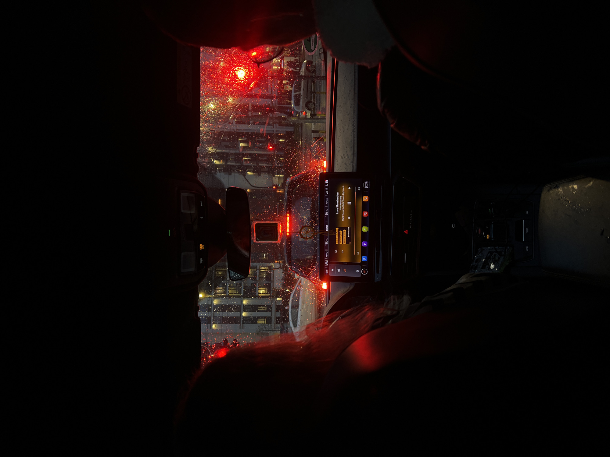 The image depicts the interior of a car, likely at night, in Belgrade, Serbia, based on the interface language and climate control readings on the car's infotainment system (16°C). It's raining, as evidenced by the water droplets visible on the windshield. The car appears to be stopped or moving slowly, as suggested by the view of city buildings, lights, and other vehicles outside the rain-streaked windshield. The driver or passenger is using the infotainment system, which displays the song "Love Illumination" by Franz Ferdinand, from the album "Right Thoughts, Right Words, Right Action." A rearview mirror is visible, reflecting the scene behind the vehicle. Red lights inside the car add to the overall dimly lit, nocturnal atmosphere.