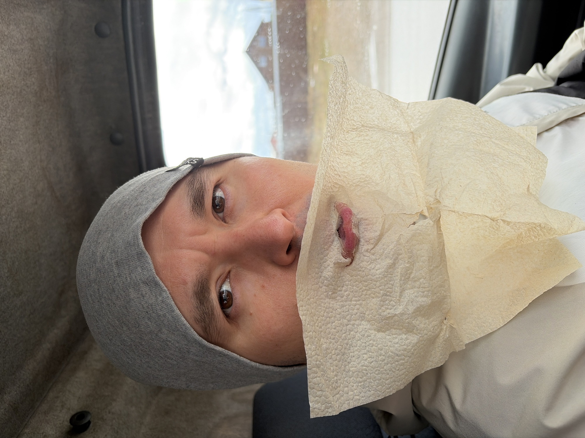 A close-up, portrait-oriented photograph shows a person wearing a gray beanie and holding a crumpled paper towel to their mouth. The person is in an automobile, as suggested by the window and interior trim visible in the background. The background outside the window is blurry but appears to show a building or structure, and indistinct features. The image is rotated, the person is facing the camera with their head tilted. The photograph's location is unknown and there is no way to derive location from the image.