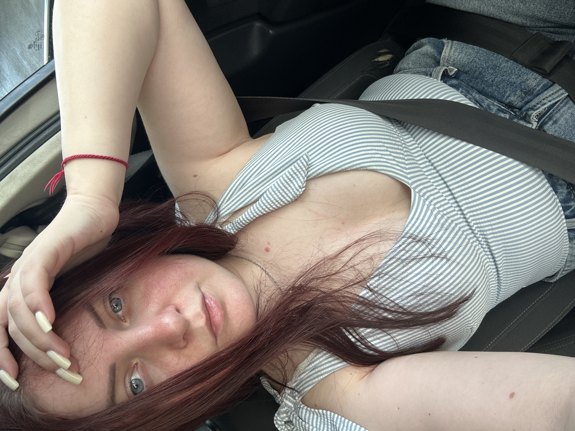 A young woman with auburn hair and blue eyes is seated in the passenger seat of a car. She is wearing a striped tank top and denim shorts, and a seatbelt is fastened across her body. Her right hand is raised to her forehead, and she is wearing a red string bracelet on her left wrist. The background appears to be the interior of a car, with dark upholstery and a window showing a blurred outdoor scene. There is no discernible indication that this image was taken in Grad Kastav, Croatia.