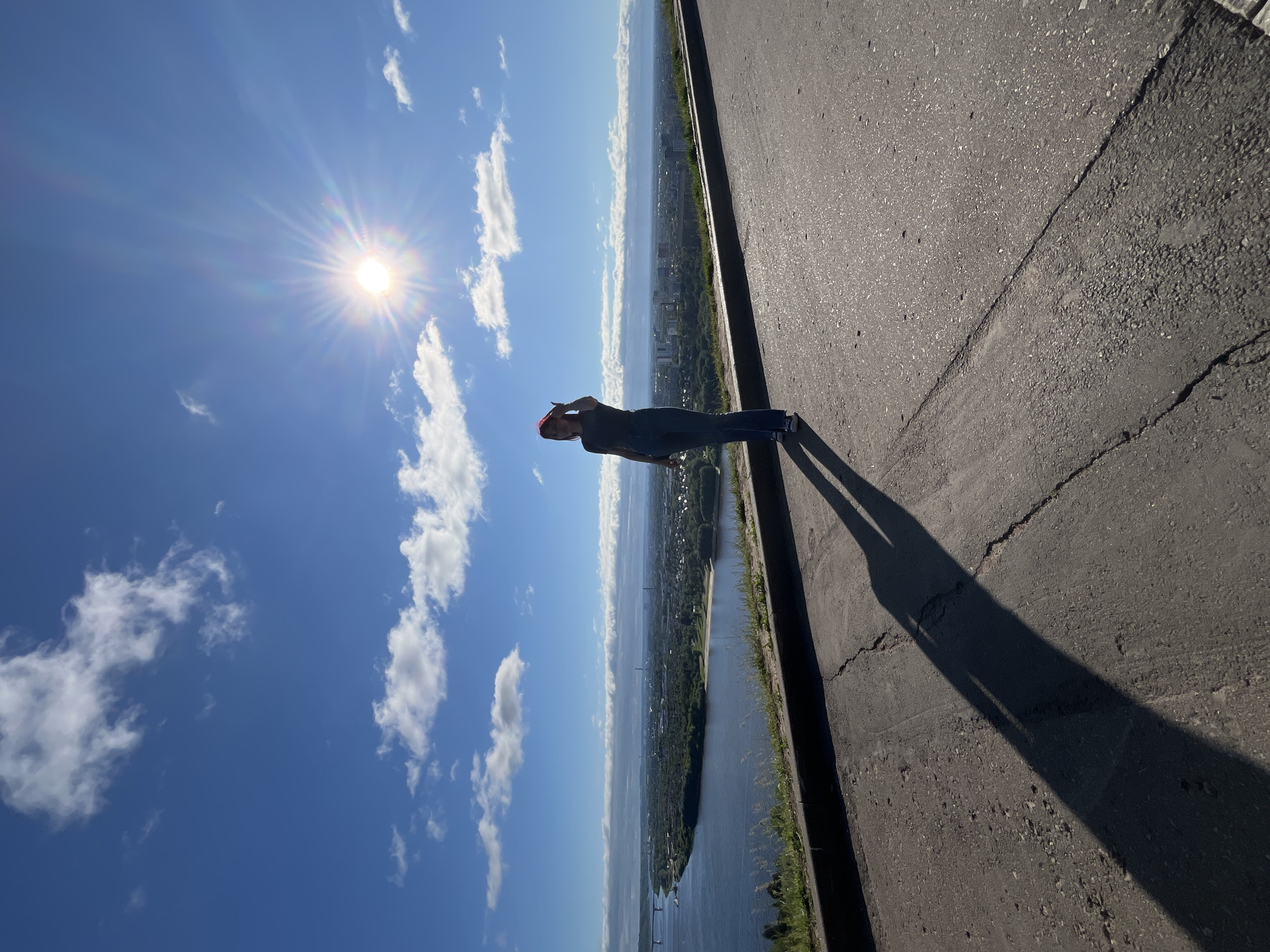 A full shot captures a person standing on a paved path overlooking the city of Nizhny Novgorod, Russia. The person, silhouetted against a bright, sun-drenched sky, is facing away from the camera and towards the city. The sun, a dazzling orb in the upper right corner, casts a long, sharp shadow of the person onto the path. The city unfolds below, a sprawl of buildings nestled beside a wide river. The sky is a vibrant blue, dotted with scattered white clouds. The paved path in the foreground shows cracks and wear, indicating it is an outdoor public space. The overall scene suggests a bright, sunny day with clear visibility over the cityscape.