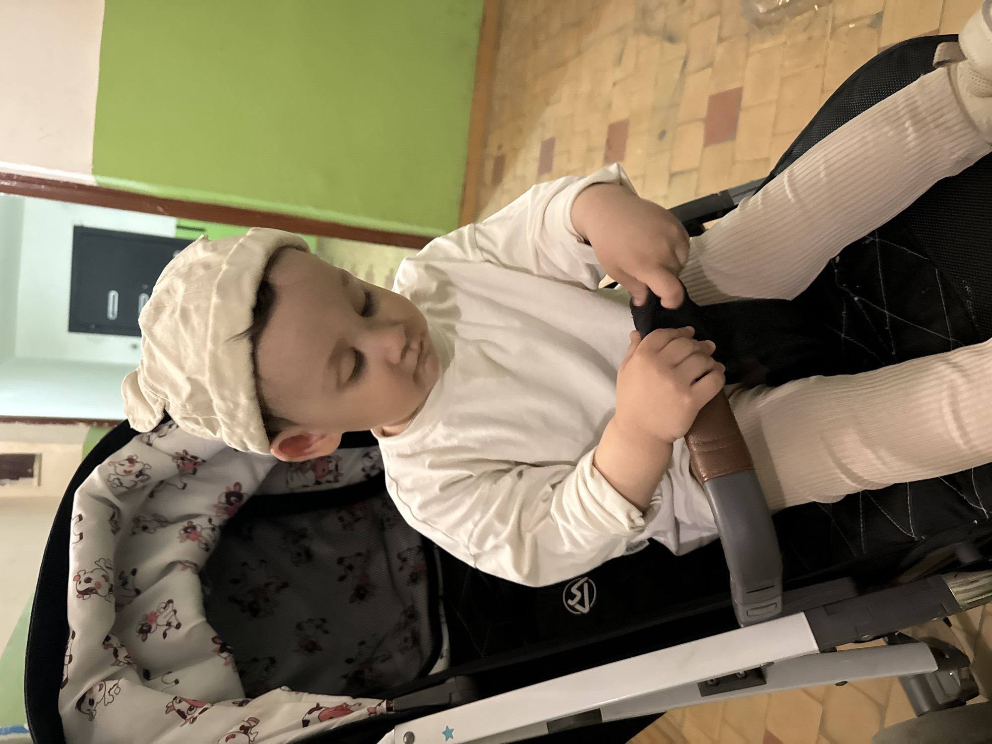 A baby is sitting in a stroller. The baby is wearing a white hat, a white long-sleeved shirt, and white leggings. The stroller is black with a white and pink cow print lining. The stroller has a white frame. The baby is holding a black strap. The background is a green wall and a tile floor. The location is indoors, and it is likely in Chaikovsky, Russia.