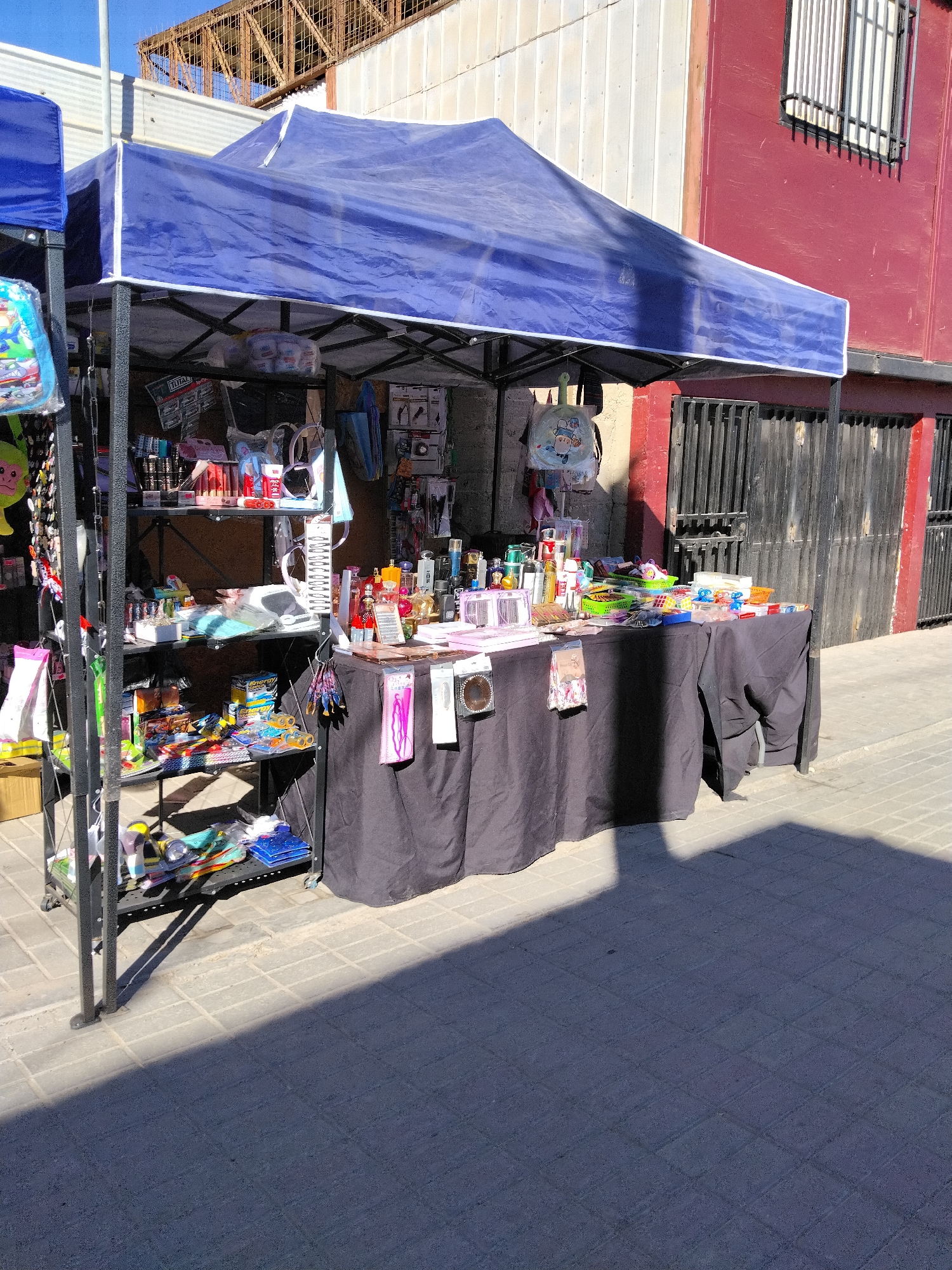 A sunny daytime view captures a roadside stall in Pozo Almonte, Chile. The stall features a blue canopy tent providing shade over a display of various goods. Underneath the tent, shelves are packed with an assortment of items, including cosmetics, perfumes, stationery, and novelty items. Some items are arranged on a black-draped table in front of the shelves. The ground is paved with interlocking bricks. In the background, a red building with metal fencing and a barred window is visible, alongside some industrial structures. The sunlight casts strong shadows, indicating it's likely midday or early afternoon. No people are visible in the scene. Visible text includes "TOTAL" on a package on one of the shelves and "ENERGY" on a box.