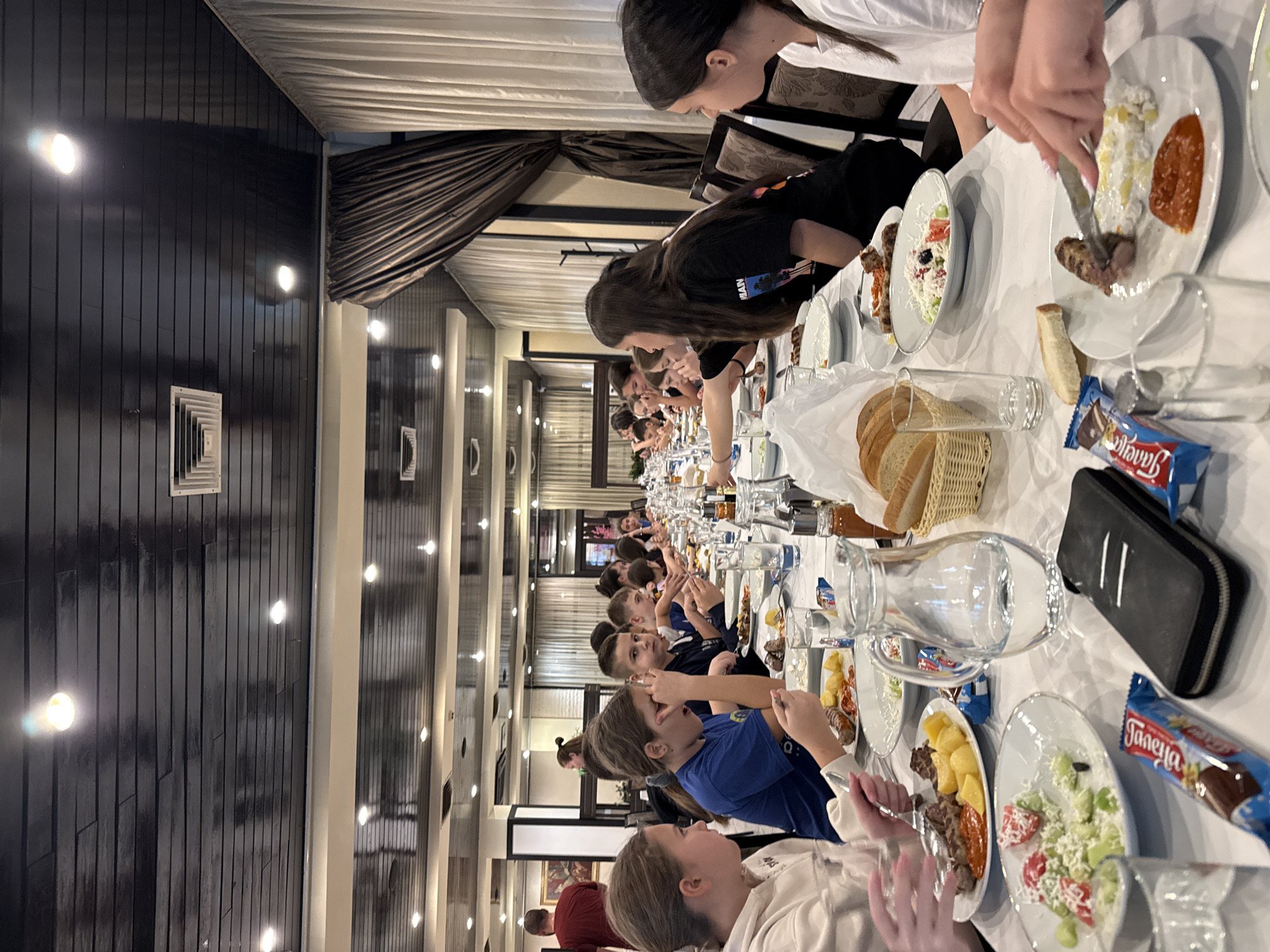 A long table is set for a large group of people. The table is covered with a white tablecloth and laden with plates of food, bread baskets, water pitchers, glasses, and candy bars. People are seated along the table, and they are eating. The people appear to be young adults and children. Some of the people are wearing blue shirts. The setting appears to be a restaurant or banquet hall. The walls are decorated with wood paneling. Recessed lighting fixtures are visible in the ceiling. The location is Etropole, Bulgaria.