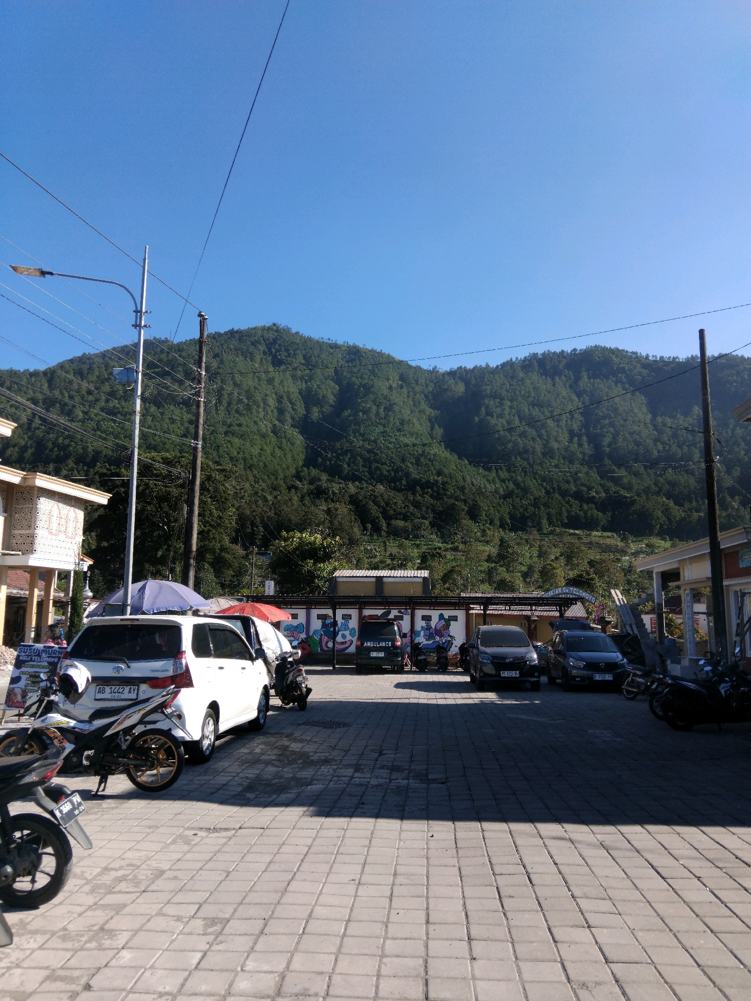 The image depicts an outdoor scene in the city of Dalangan, Indonesia, on a clear, sunny day with a bright blue sky. In the background, a densely forested mountain range dominates the landscape, with terraced fields visible on its slopes. In the foreground, a paved area, likely a parking lot, is lined with gray interlocking concrete pavers.
Several vehicles are parked in the lot. A white van is prominent in the center-left, with a license plate reading "AB 1442 AY". To its right, two dark-colored cars are parked, one with a license plate starting with "AE" and the other with "B". A motorcycle is parked behind the white van, and another motorcycle is visible on the far left. Two motorcycles are also parked on the right side of the image.
Buildings line the right and left sides of the frame. The building on the left appears to be a small commercial establishment with decorative patterned walls. The buildings on the right seem to be part of a complex, with one structure featuring a light-colored roof and a wall with graffiti. A street lamp is mounted on a pole on the left side of the image, and utility poles with numerous wires are visible throughout the scene. Two umbrellas, one blue and one red, are set up in the parking area. A sign with Indonesian text is partially visible on the left. The image captures a moment in a public space, likely a market or a rest stop, with vehicles indicating recent activity.