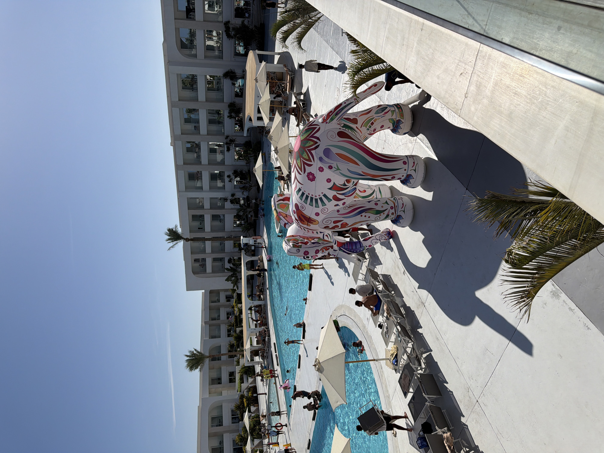 The image shows a resort swimming pool area in Sharm El Sheikh, Egypt. The pool is large and blue, with several people swimming and lounging around it. There are lounge chairs and umbrellas scattered around the pool deck. In the foreground, there is a large, colorful inflatable elephant. The resort buildings are modern and white, with balconies overlooking the pool area. Palm trees are visible throughout the scene. The sky is clear and blue.