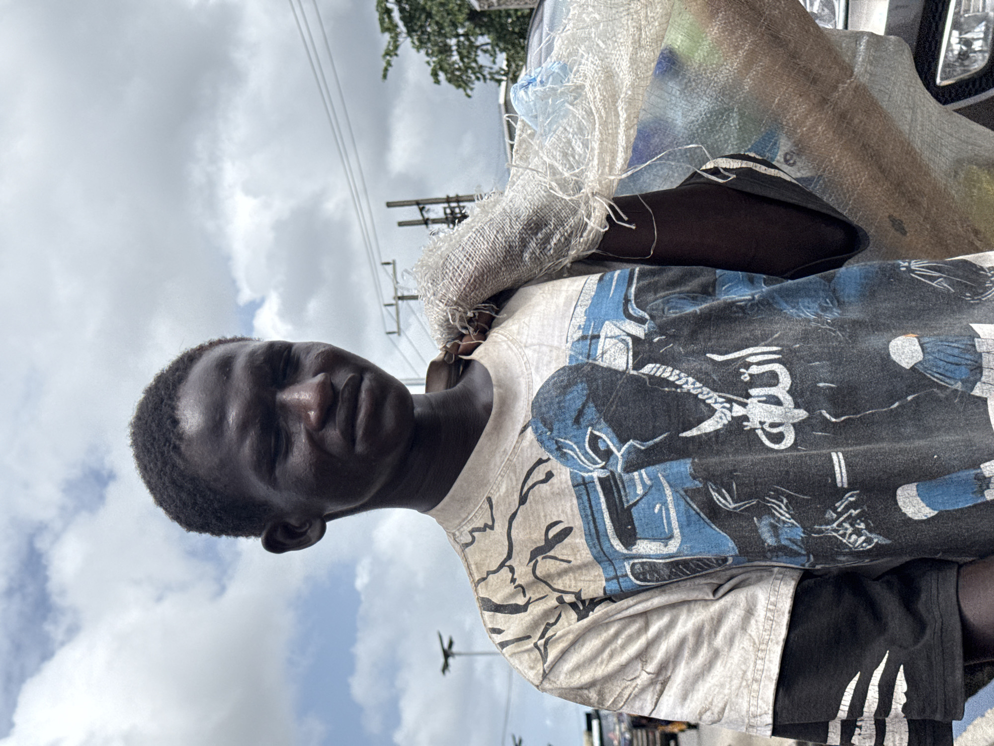 A young man is standing in Ado, Nigeria, wearing a patterned t-shirt and carrying a white sack on his shoulder. The sky is cloudy. Power lines are visible in the background. A vehicle is partially visible on the right side of the frame.