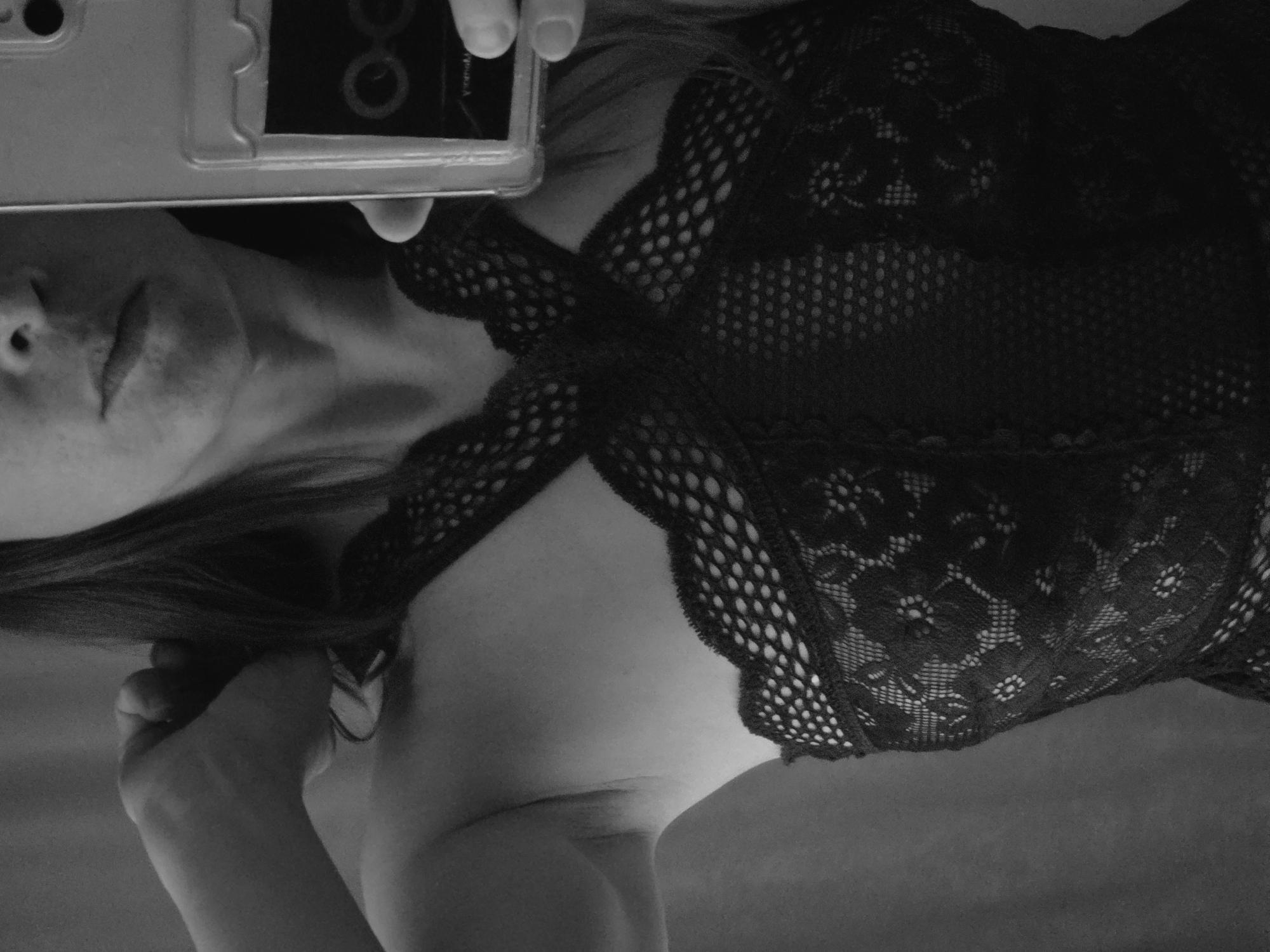 A black and white, vertically oriented photograph captures a partial view of a person, likely an adult female, in an intimate, indoor setting, appearing to take a selfie. The subject is wearing a black, intricately designed garment, possibly lingerie or a bodysuit, featuring distinct lace and mesh patterns across the upper torso.
The person's head is positioned on the left side of the frame, tilted with eyes closed or looking downwards, and lips softly closed. Long, dark hair falls over their left shoulder. Their left hand, with visible thumb and fingers, firmly holds a smartphone encased in clear plastic in the upper left corner of the image. The phone's screen shows a dark background with a prominent light circular icon and faint text that appears to be "Veronica" or similar lettering.
The focus is largely on the upper body, showcasing the detailed textures of the lace and mesh fabric. The lace exhibits floral or geometric patterns, while the mesh sections reveal glimpses of skin underneath. Another hand is visible on the right side of the frame, resting gently against the person's side, below the chest. The lighting is soft, creating subtle shadows and highlights that accentuate the contours of the body and the fabric. The background is uniformly dark and indistinct, suggesting a private, interior space. The overall mood of the image is intimate and self-reflective.