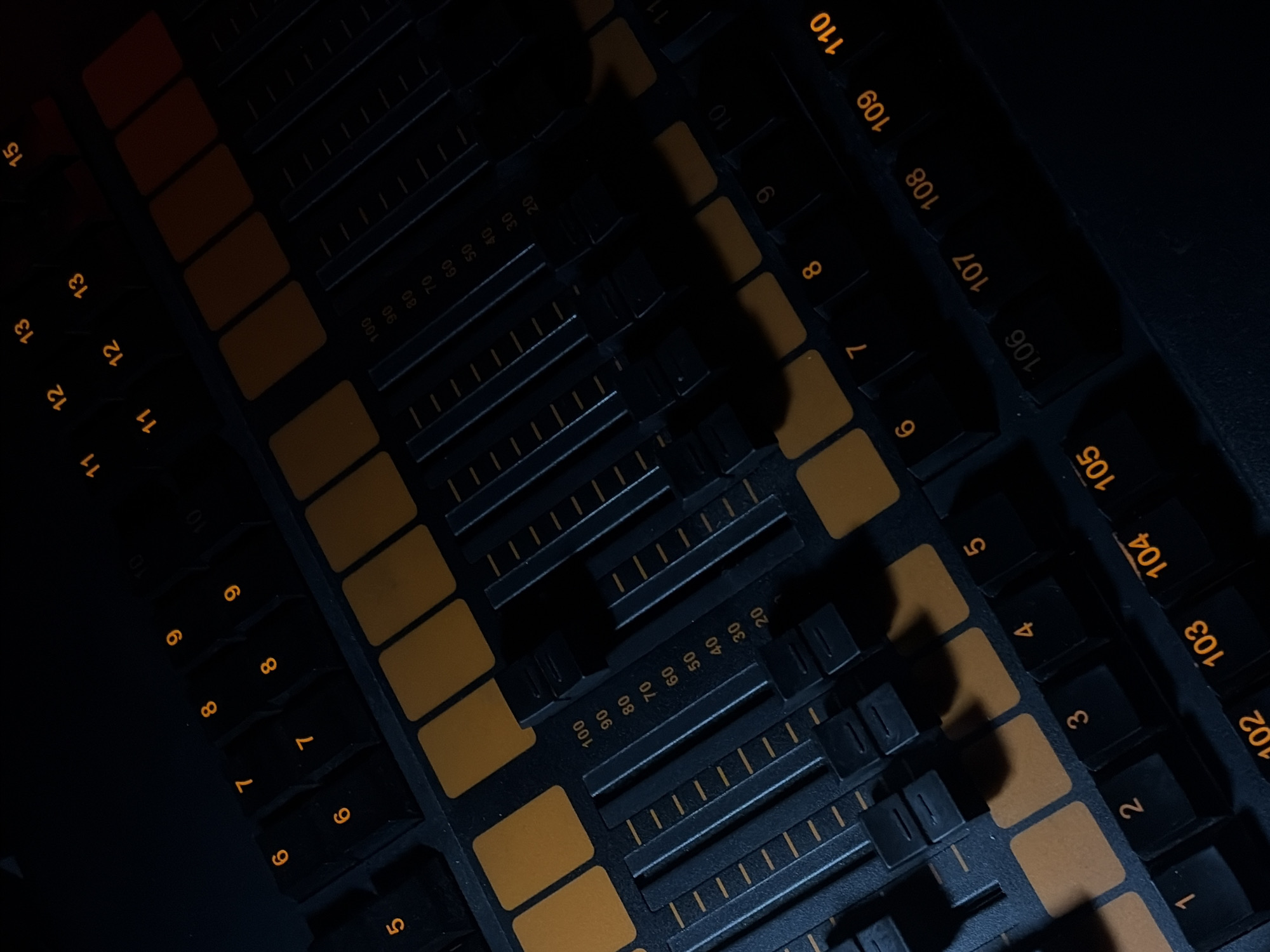 The image displays a close-up, angled view of a dark-colored control console, likely for audio or lighting, within a dimly lit indoor environment. The console features numerous black square buttons illuminated with glowing orange numbers, including sequences like 1-9, 11-15, and 102-110. Multiple vertical fader tracks are visible, each marked with a numerical scale from 20 to 100. Several solid orange rectangular buttons are integrated into the control surface. No individuals are present, and no actions or interactions are depicted. The environment offers no discernible details regarding its location in the city of Moscow, Russia.