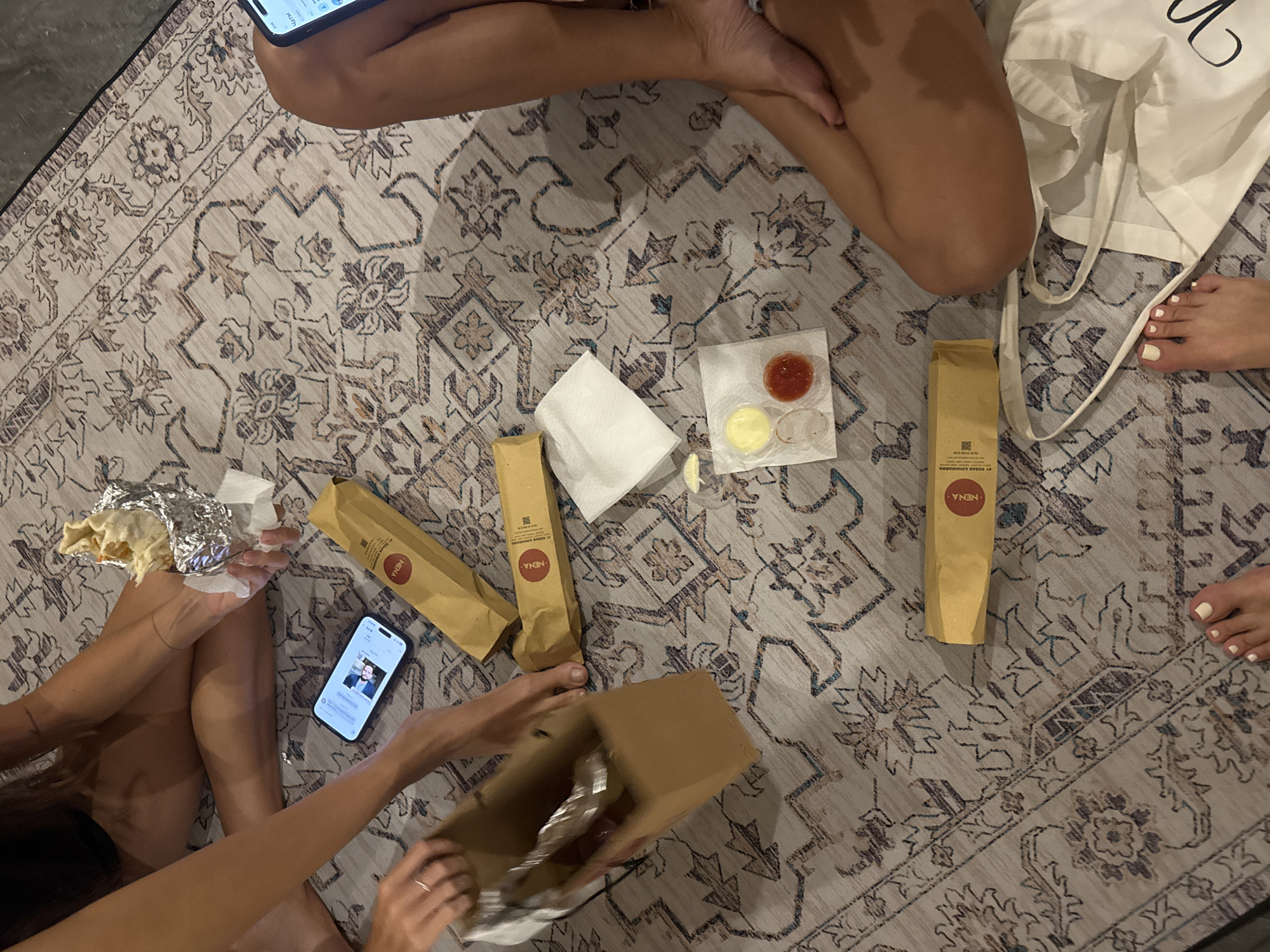 A group of people is sitting on a rug, seemingly indoors. They are surrounded by takeout food from "NENA: ST. KEBAB & SHAHARAMA." The food includes wrapped sandwiches, sauces, and a cardboard box with foil-wrapped items. One person is looking at a phone, and others are reaching for the food. The rug is patterned, and the environment seems casual. The location is in North Kuta, Indonesia.