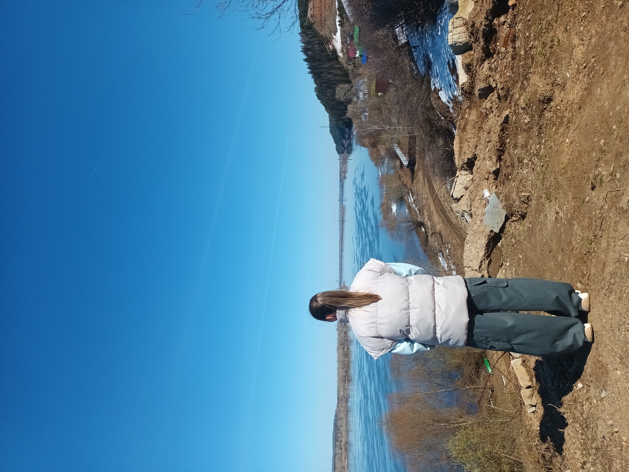 A person stands on a dirt cliff overlooking a river in Ольховка, Russia. The person wears a light-colored puffy jacket and dark pants. The river has some ice, and there are trees on the other side. The sky is blue.