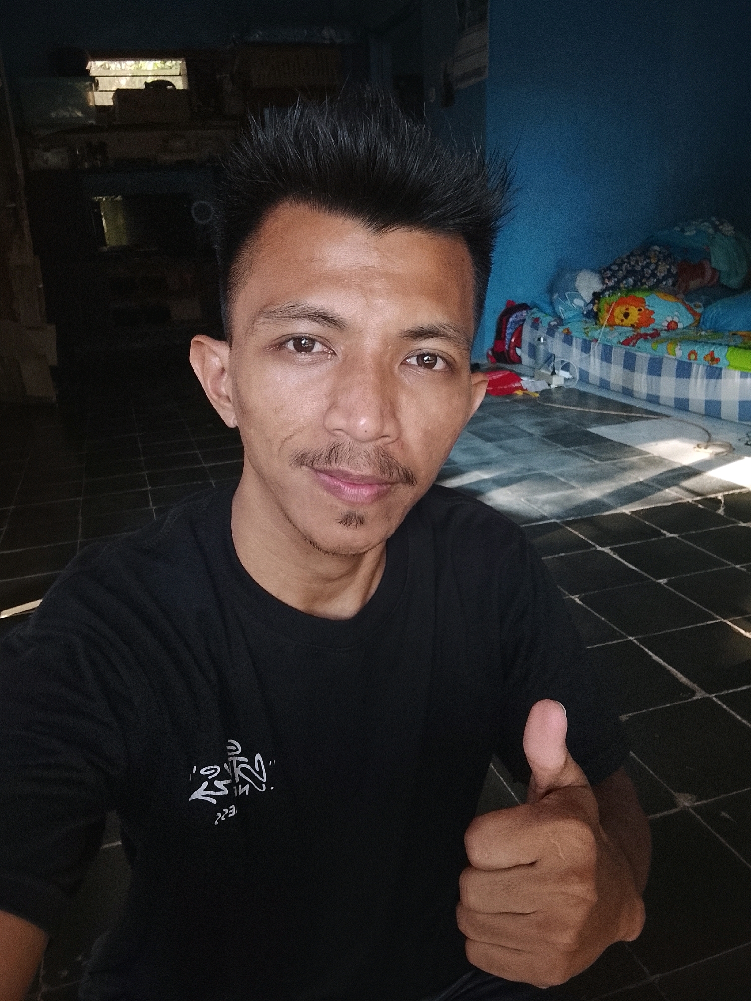 A man is shown in a selfie, giving a thumbs-up. He is wearing a black t-shirt with white writing on it, and has spiky black hair. He is standing in what appears to be a living room, with a tiled floor and a blue wall. There is a bed with a checkered blue and white comforter and colorful pillows in the background. The scene is dimly lit, with sunlight streaming through a window and casting shadows on the floor. The man is looking directly at the camera with a slight smile. The photo is taken from a slightly low angle.