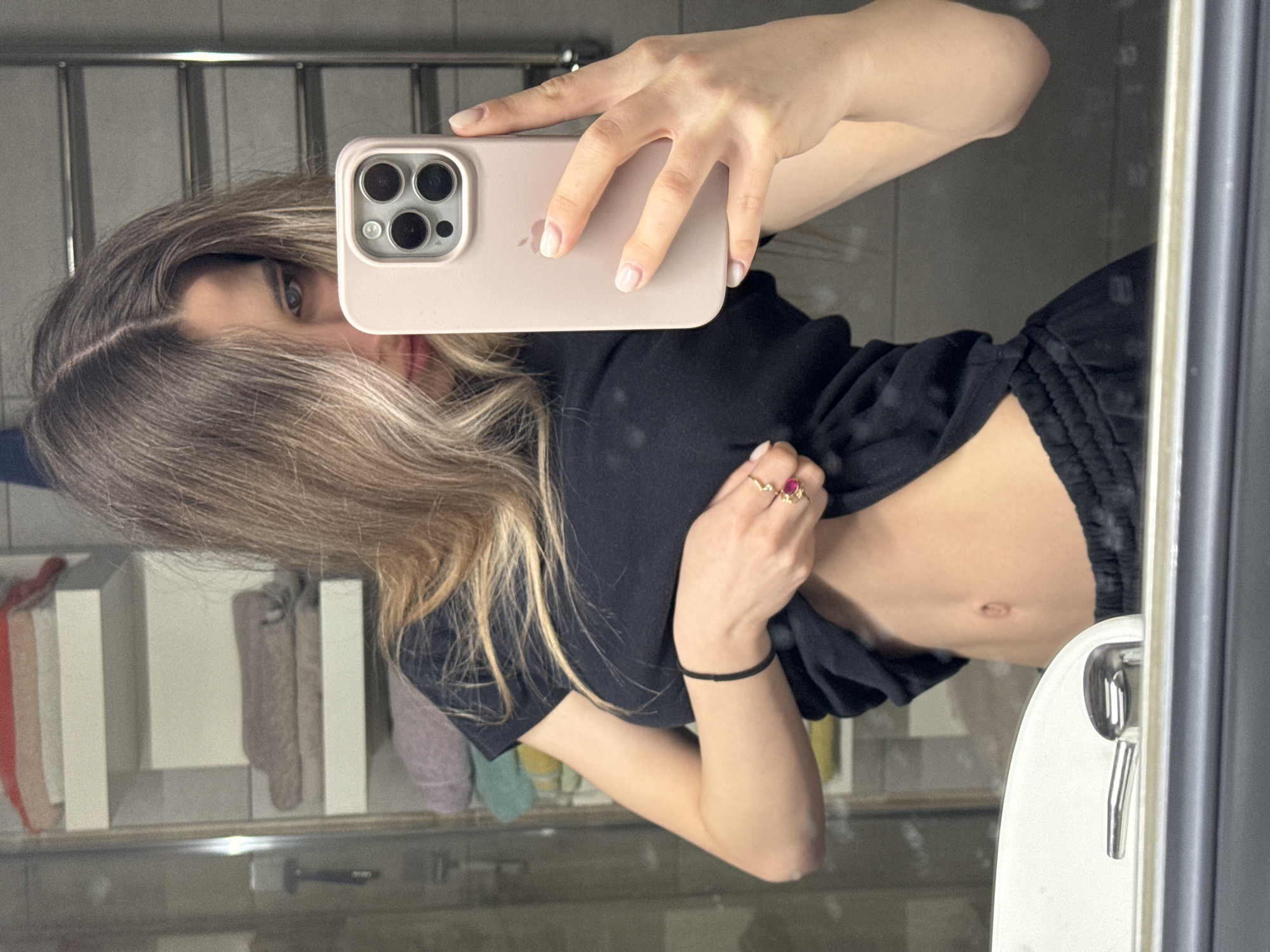 A mirrored selfie appears to be taken in a bathroom. A woman is holding a pink-cased iPhone, partially obscuring her face. She is wearing a black T-shirt and black sweatpants, revealing a portion of her abdomen. She has light brown hair with dark roots. Her left hand is visible, adorned with a ring and a black wristband. The background includes towel racks, towels, and a sink. The location is not definitively discernible as Chaikovsky, Russia, based solely on visual information.