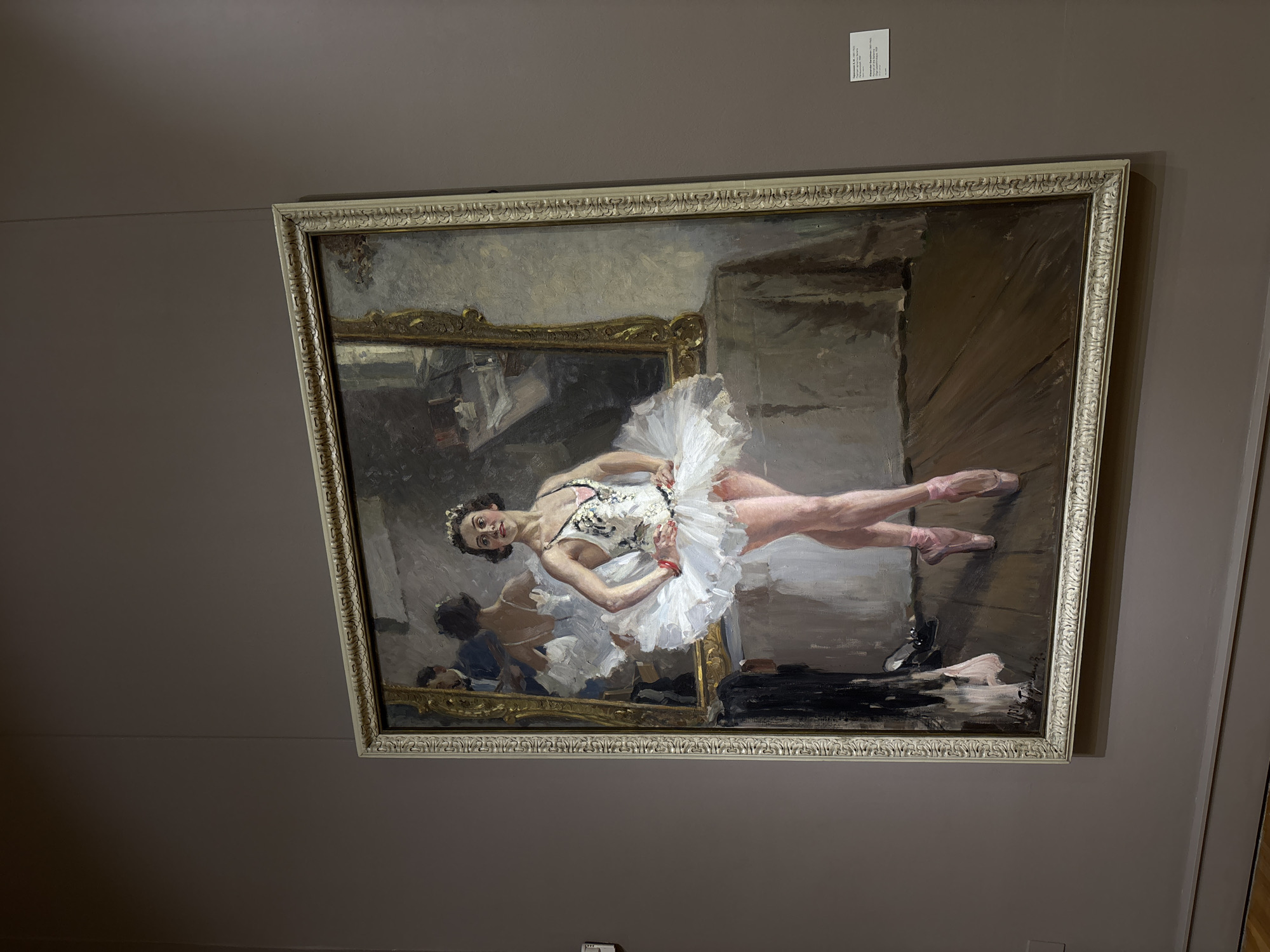 A framed painting hangs on a wall in a museum. The painting depicts a ballerina in a white tutu stretching in front of a mirror. The mirror reflects a second ballerina. The painting is of a muted color palette, dominated by creams, browns, and grays. The frame is ornate and pale. A small white placard is affixed to the wall above the painting. The wall is painted in a neutral gray tone. A section of a door frame is visible on the right. The painting is "Rehearsal on Stage" by Edgar Degas, which hangs in the Pushkin State Museum of Fine Arts in Moscow, Russia.