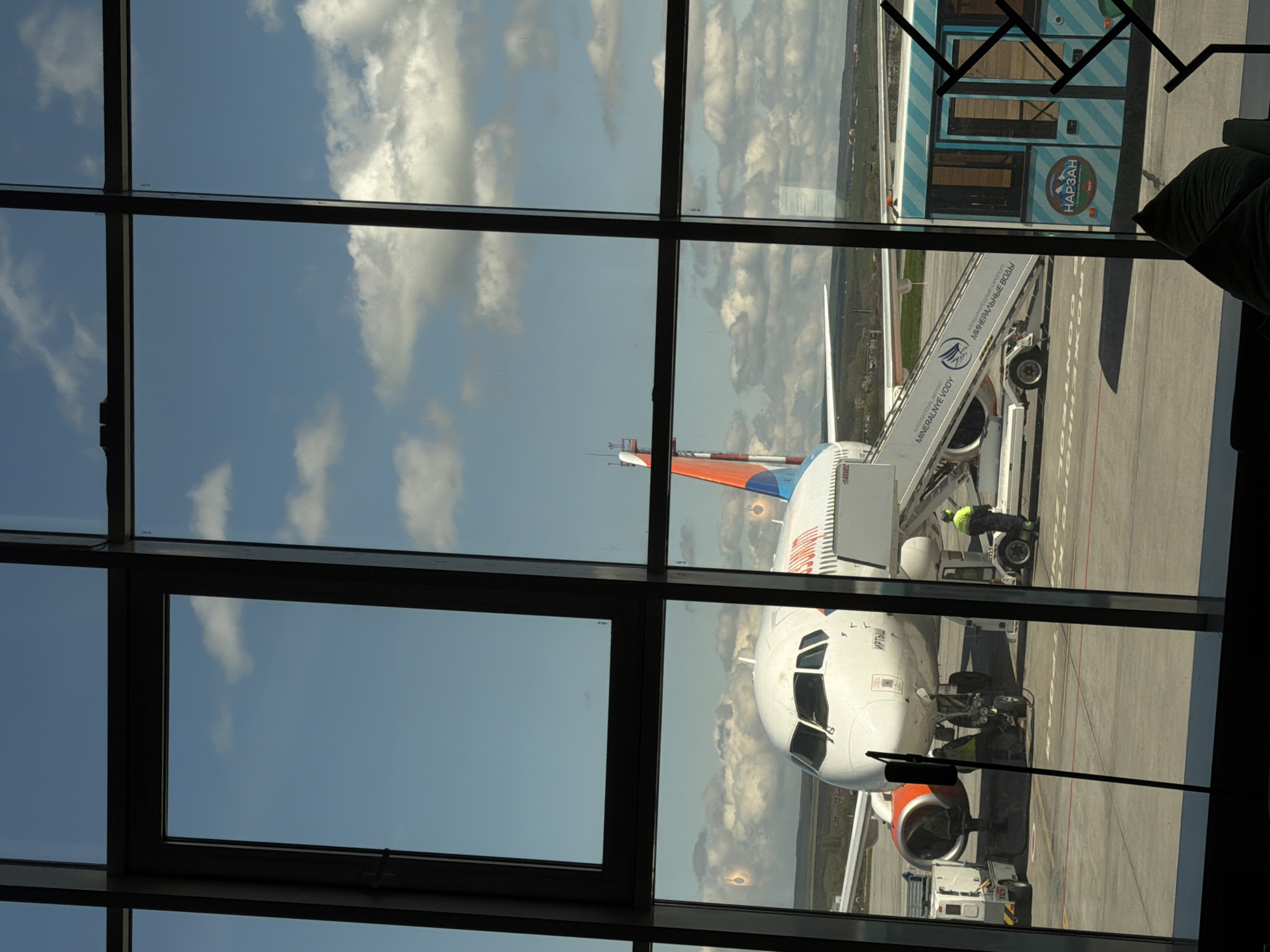 The image captures an outdoor scene at an airport, viewed through a multi-pane window from inside a terminal building. An airplane, predominantly white with orange and blue accents on its tail and engine nacelles, is parked on the tarmac. Its nose, bearing the Cyrillic name "ИРТЫШ" (Irtysh), is visible in the lower right section of the frame.
A set of mobile passenger stairs is connected to the aircraft's forward door. On the side of these stairs, text in Cyrillic reads "ВАНЬКОВЫЙ ПРОЦЕСС МИНЕРАЛЬНЫЕ ВОДЫ" and "MINERALNYE VODY", accompanied by a logo resembling a bird or eagle. Several ground crew members are present around the aircraft; one individual in high-visibility yellow attire is near the front of the plane by the stairs, while another is visible near the front landing gear.
To the right of the airplane, a light blue and white striped bus or shuttle is parked. It features the Cyrillic word "НАРЗАН" (Narzan) with a mountain-themed logo, and also "АЭРОПОРТ" (Airport). The tarmac has markings, including partially visible Cyrillic text "КОНТРОЛЬ" (Control).
The sky, filled with scattered white clouds, is visible through the window panes, and reflections of these clouds are also noticeable on the glass, creating a sense of depth. The day appears bright and sunny. The dark frames of the window panes divide the scene into multiple rectangular sections. A person's arm or shoulder is faintly visible in the extreme right, suggesting someone is observing the scene. The location, Загорский, Russia, is consistent with the Cyrillic text observed.