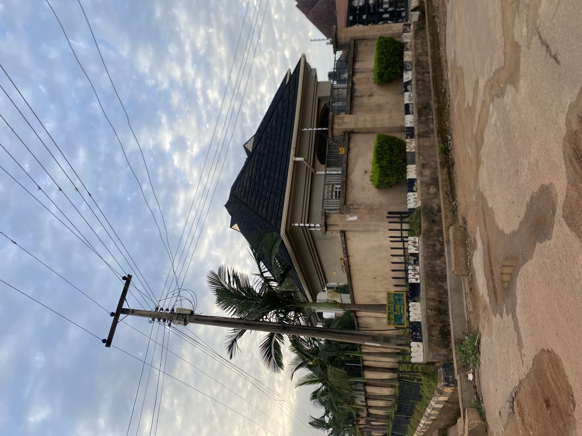 This image depicts a street scene in Bar Jirgi Summa, Nigeria, dominated by a residential building and its surrounding fence. The building features a dark, multi-angled roof and is painted a light beige color. It is partially obscured by a high fence made of stone and concrete, topped with security wiring. Two neatly trimmed green bushes flank the entrance to the property.
In front of the house, a row of tall palm trees runs along the perimeter. A prominent wooden utility pole, laden with electrical wires, stands to the left of the house, partially in front of the palm trees. A street sign with green lettering on a yellow background is visible on the pole, reading "YAKUBU CHOJI-DAVOU CLOSE" with an arrow pointing to the right.
The street in the foreground is paved with an uneven surface, showing patches of dirt and puddles of water, suggesting recent rain. The sky above is partly cloudy with a mix of blue and white clouds, indicating diffused daylight. The overall impression is that of a residential neighborhood in a tropical or sub-tropical climate. There are no people visible in the frame.