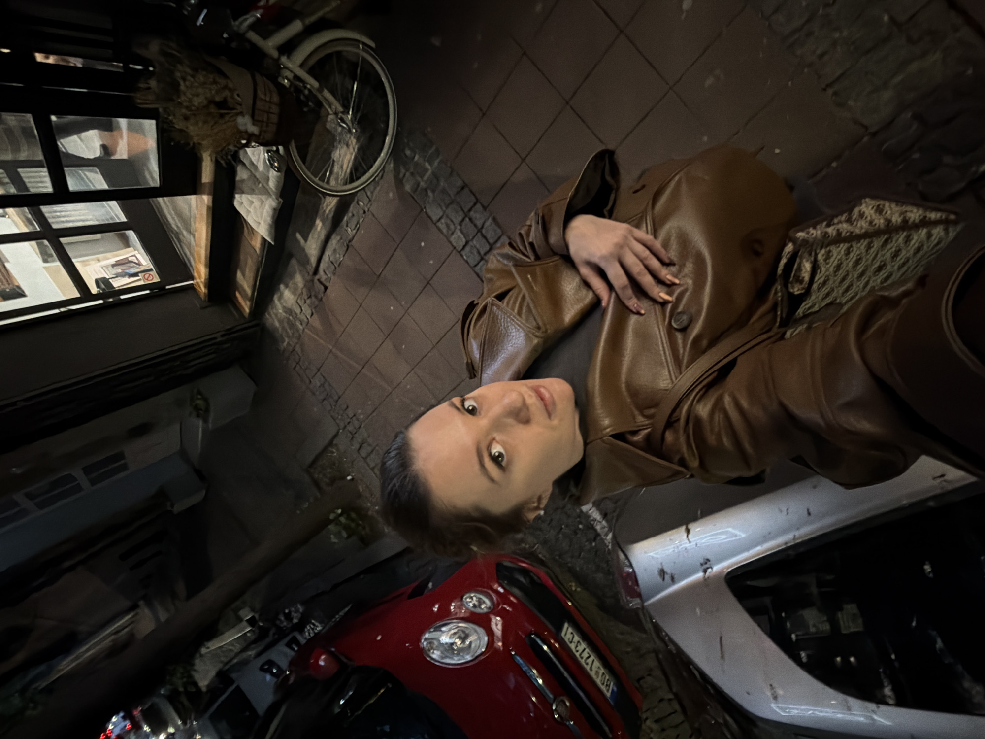 The image, oriented correctly, captures a night-time street scene in an urban European setting. The main subject is a young woman, seen from the chest up, positioned slightly to the left of the frame. She has medium-brown hair, pulled back, and is looking directly at the camera with a neutral to slightly serious expression. She is wearing a brown, possibly leather, trench coat, and her left hand with manicured, light-colored nails rests on her coat. A patterned, woven bag is visible behind her right shoulder.
The background reveals a cobblestone street or pavement, suggesting an older part of a city. On the left side, a bicycle with a white frame and wheels is parked or hanging against a dark building facade. The building has windows reflecting interior lights and details, including a faint "No Smoking" sign. On the right and lower part of the image, two cars are partially visible. A red car, resembling a Fiat 500, occupies the lower center, with its front grille and headlights visible. Its license plate clearly displays "BG 1273-CI," with "BG" being a common abbreviation for Belgrade, Serbia. To the right of the red car, the silver rear bumper and trunk area of another car are seen, appearing somewhat dusty or dirty.
The scene is dimly lit, indicative of night, with light sources primarily coming from within buildings and reflections on surfaces. The woman appears to be posing for a photo, likely a selfie.