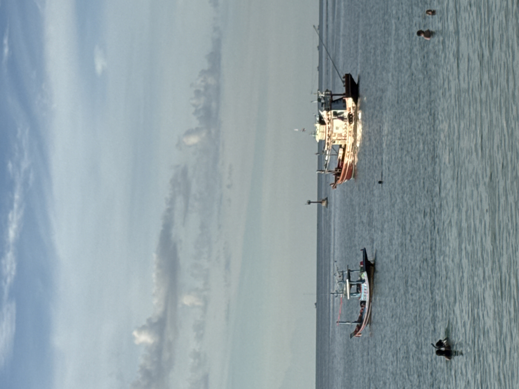 This image captures a serene seascape near Koh Phangan, Thailand. In the foreground, two individuals are partially submerged in the water, one wearing a snorkel mask. Further out at sea, a smaller fishing boat, characterized by its red and white hull and a blue canopy, is anchored. A larger, more ornate fishing vessel, with a prominent white cabin and red trim, sails in the mid-ground. In the background, a lighthouse-like structure stands on a buoy in the water, and the horizon is dotted with a few distant boats and ships. The sky above is a pale blue, adorned with scattered white clouds, suggesting a pleasant day with calm waters. The overall scene conveys a sense of tranquility and outdoor leisure.