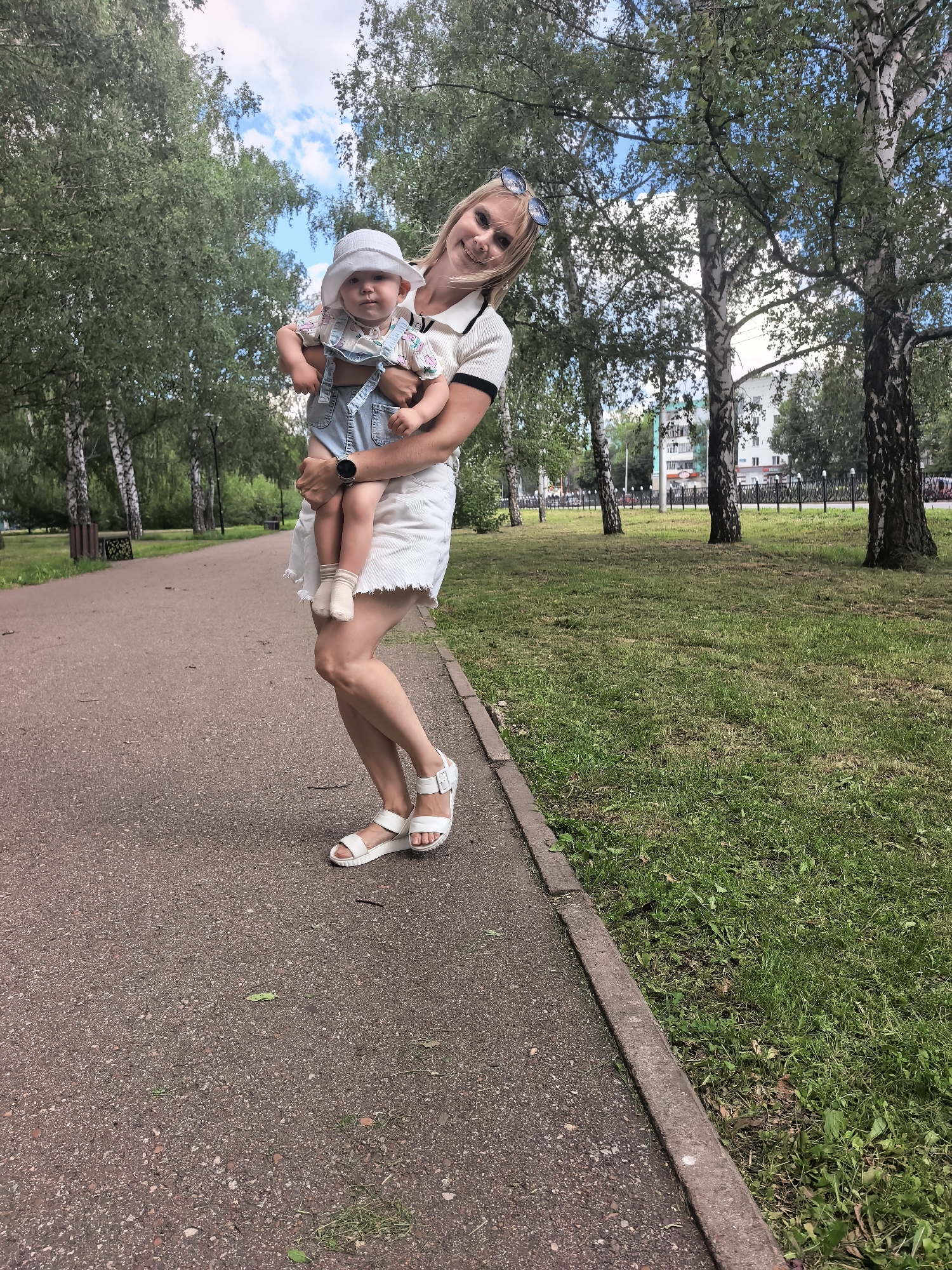 A blond woman in a white skirt and sandals holds a baby wearing a white hat and denim overalls in a park. The woman is smiling and looking at the camera, while the baby is looking forward. The park features birch trees, a paved walkway, and a grassy area. Buildings can be seen in the background. The scene appears to be in the city of Sterlitamak, Russia.