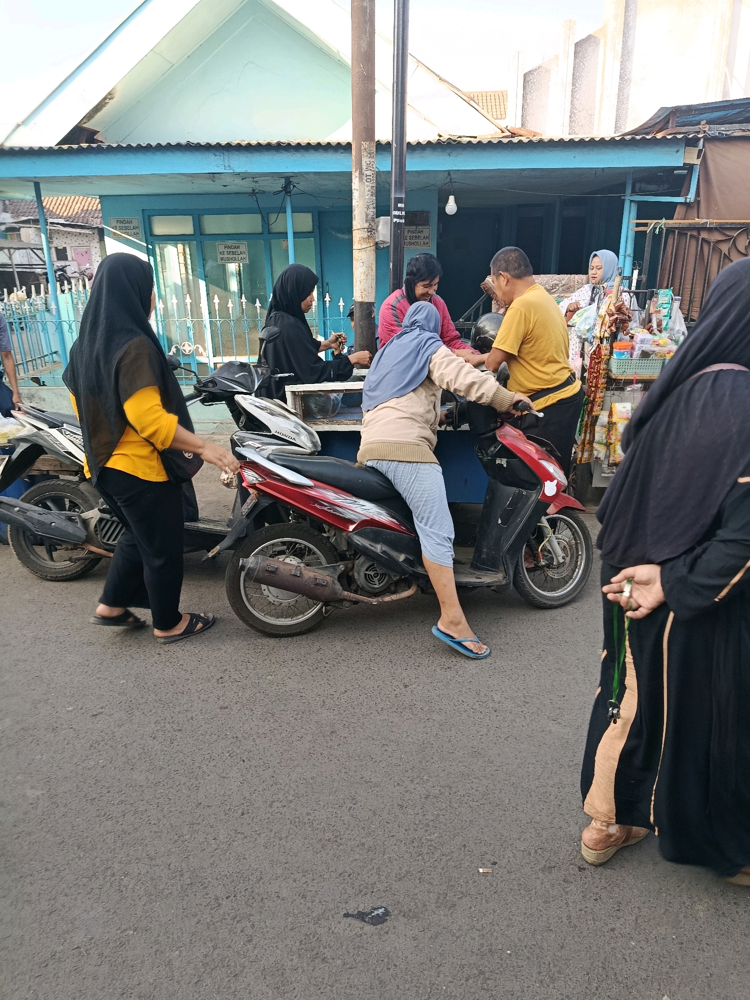 In Gondanglegi, Indonesia, a street scene unfolds with several individuals and motorcycles. A woman wearing a black hijab and a yellow long-sleeved shirt is standing next to a motorcycle, with her left hand resting on its handlebars. To her right, another woman in a light brown hijab and pants is seated on a red motorcycle, adjusting something on its controls. A man in a yellow t-shirt is leaning over the motorcycle, facing the seated woman. In the background, two other women in black hijabs are visible, one in front of a blue building with signs, and another further back near a stall displaying various items. A third motorcycle is parked to the left of the woman in the yellow shirt. The environment suggests a typical street setting with a building and pavement.