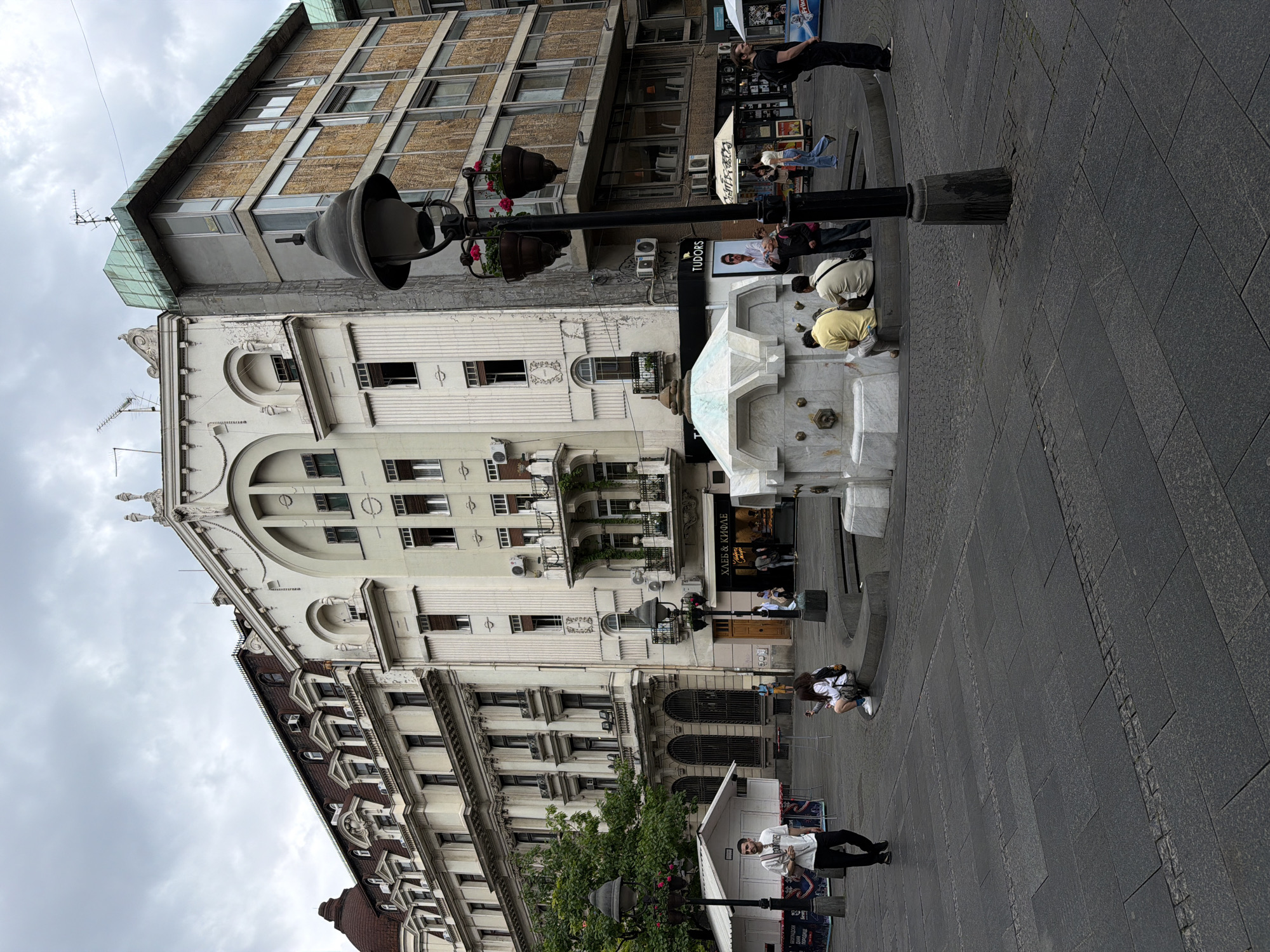 The image depicts a street scene in Belgrade, Serbia. The primary focus is on an ornate fountain in a public square, with people interacting with it. Some individuals are leaning over the fountain, seemingly looking or interacting with the water. In the background, there are several buildings with distinct architectural styles, including a mix of older, ornate facades and more modern structures. A black lamp post with flower boxes is also visible. The ground is paved with stone, and there are other people walking or standing in the background. The sky is overcast, suggesting a cloudy day.