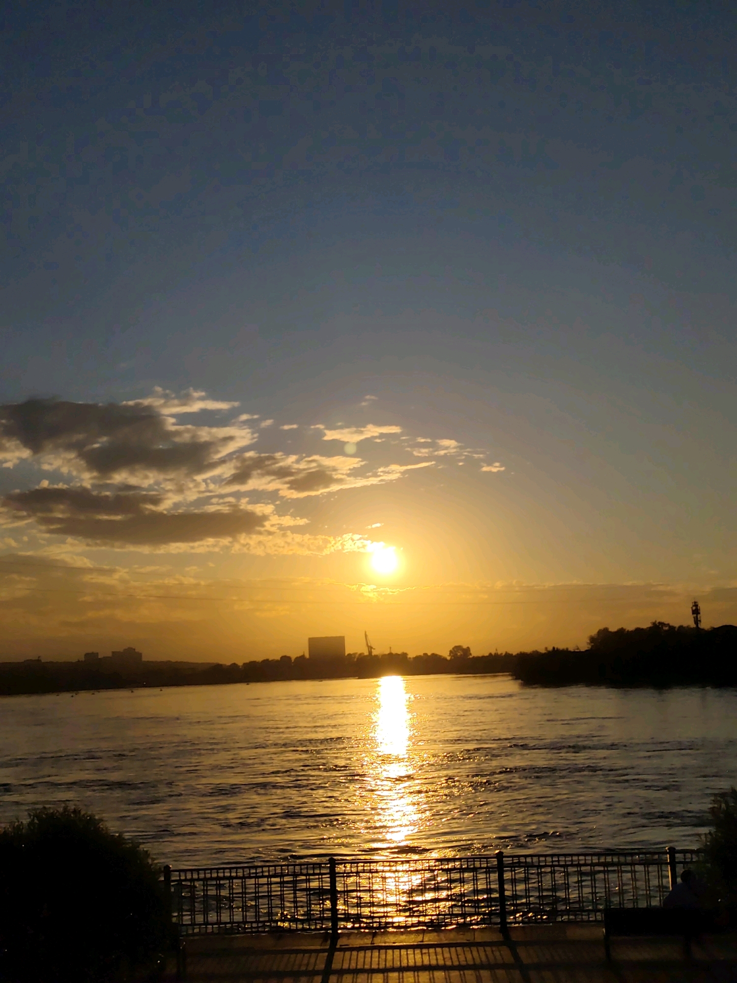 The image depicts a sunset over the Angara River in Irkutsk, Russia. The sun is low on the horizon, casting a golden glow across the water and the sky. Wispy clouds are scattered across the upper part of the frame, tinged with orange and yellow. In the distance, the silhouette of the city skyline is visible, with buildings and industrial structures. The foreground features a dark railing and a bench, suggesting a public viewing area along the riverbank. The water is rippled, reflecting the vibrant colors of the sunset. The overall scene is serene and picturesque, capturing the beauty of the natural landscape at dusk.