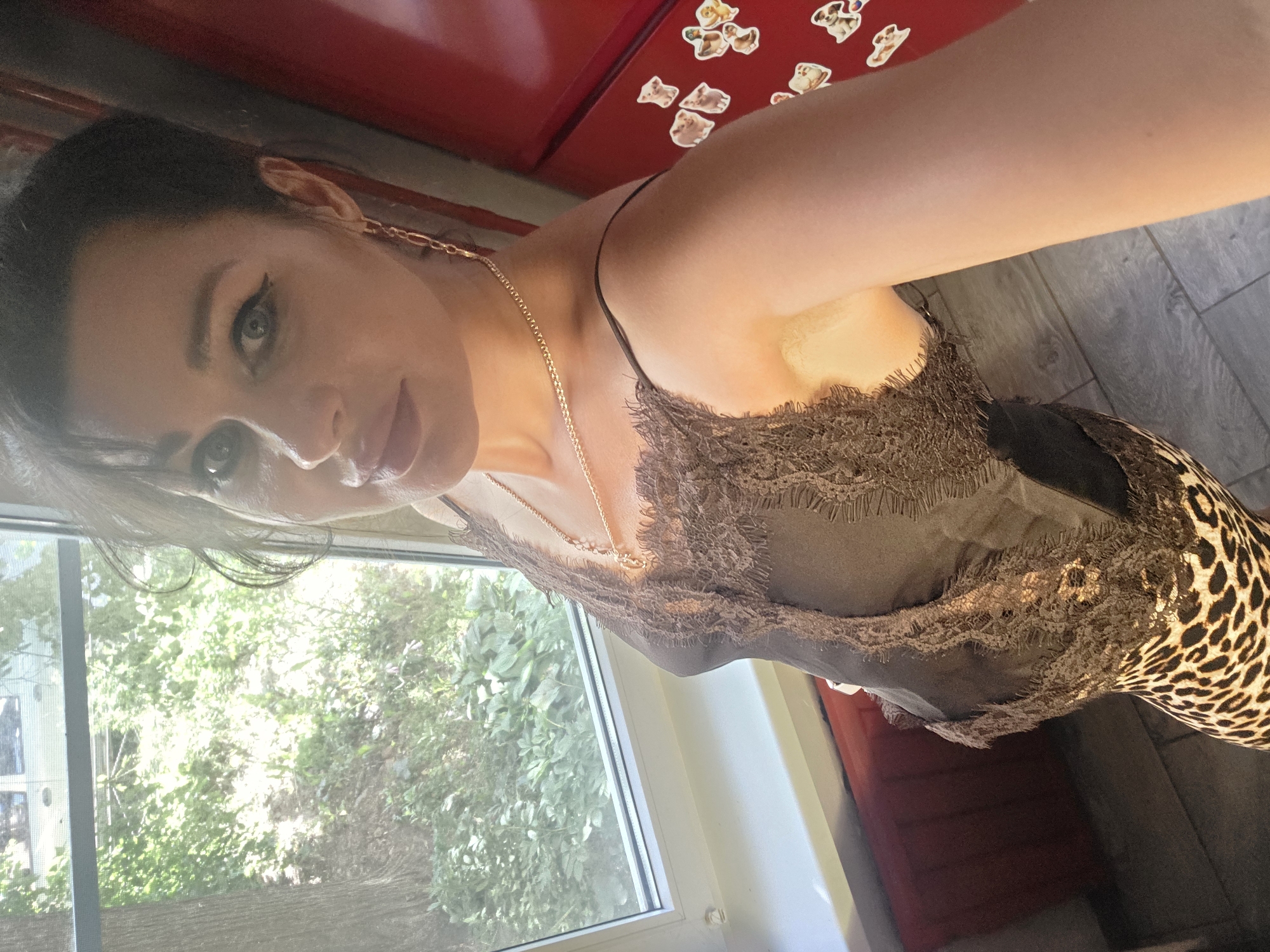 The image depicts a woman posing for a selfie in what appears to be an indoor setting. She is wearing a dark, lacy top and a leopard-print skirt. The background includes a window with greenery visible outside, suggesting a residential or apartment building. A red appliance, possibly a refrigerator, is visible on the right side of the frame, adorned with stickers. A radiator is also present beneath the window. The woman has dark hair styled up and is wearing earrings and a necklace. Her makeup includes winged eyeliner and noticeable lip color. The lighting suggests a daytime setting, possibly with natural light coming from the window. The floor appears to be tiled. There is no indication of the specific location within Saint Petersburg, Russia.
