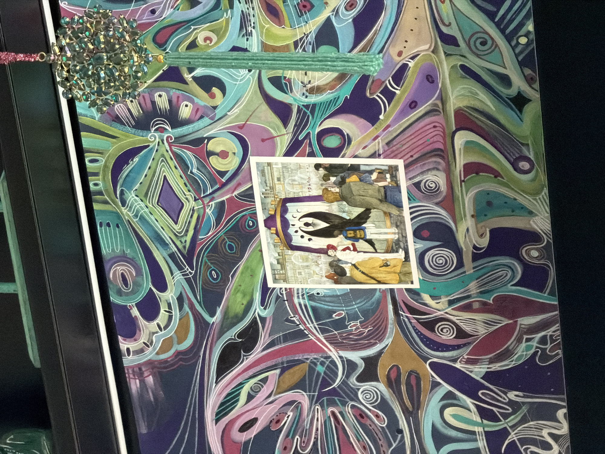 A close-up, slightly angled shot reveals an abstract artwork affixed to a dark-framed panel. The artwork, predominantly featuring swirls and patterns in shades of purple, blue, green, and pink, has a chaotic yet organic feel. A textured, light green horizontal element rests across the upper middle section of the painting. To the left of this, a large, circular decorative piece covered in small gems is attached with what appears to be a pink, glittery string.
Positioned centrally on the artwork is a small, rectangular print. This print depicts an illustration featuring human figures. The style is detailed, suggesting a historical or fantasy theme.
The background is dark, providing contrast and highlighting the artwork. The image has a decorative, artistic, and somewhat whimsical quality. There is no evidence in the image to indicate this is in Moscow.