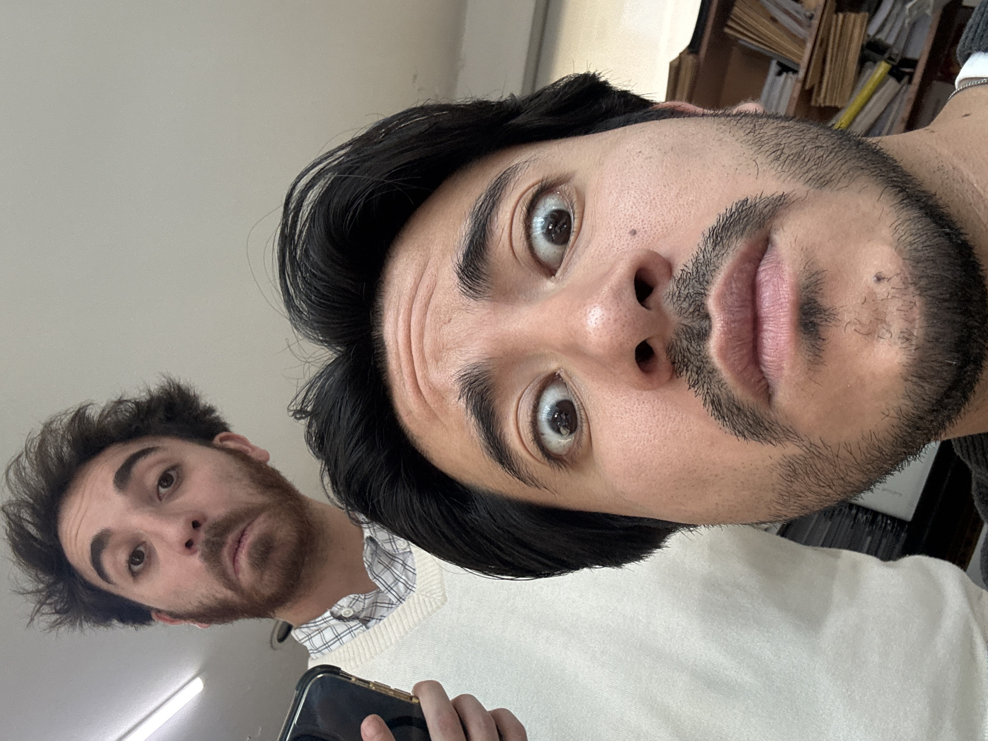 Two men are visible in the image, both likely in their late 20s or early 30s. The man closer to the camera has dark hair and a full beard. He appears surprised, his eyes wide. The second man, further back, also has dark hair and a beard. He is holding a black phone. The background is indistinct but suggests an interior setting, possibly in Puebla, Mexico, given the request context, with the possibility of furniture or shelves in the distant background. The lighting is adequate, and the camera angle is slightly upward.