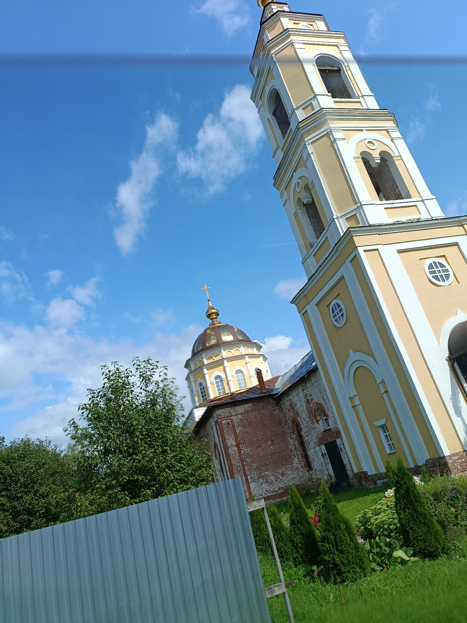 The image shows a bell tower and a church dome against a blue sky with white clouds. A corrugated metal fence runs along the bottom of the frame, and there are trees and manicured bushes in the foreground. The bell tower is painted yellow with white trim and has arched openings. The church dome is golden and topped with a cross. The church building itself is made of red brick with white architectural details. The scene appears to be located in the village of Pashnevo, Russia, judging by the architectural style and the overall setting. There are no people visible in the image.