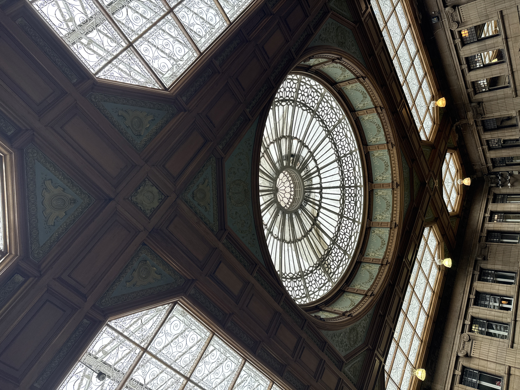This is a low-angle, full shot of the interior of the Scottish National Gallery in Edinburgh, United Kingdom, looking up at the ornate ceiling and a domed skylight. The ceiling is a complex structure of dark wood panels with decorative teal and gold friezes. The central focus is a large, circular skylight with a radiating pattern of glass panes and metalwork, surrounded by a decorative band of figures and foliage. Flanking the central dome are smaller, rectangular skylights, also featuring intricate metalwork. Below the skylights, the walls of the gallery are visible, adorned with classical architectural details and rows of windows. The overall impression is one of grandeur and historical architecture.