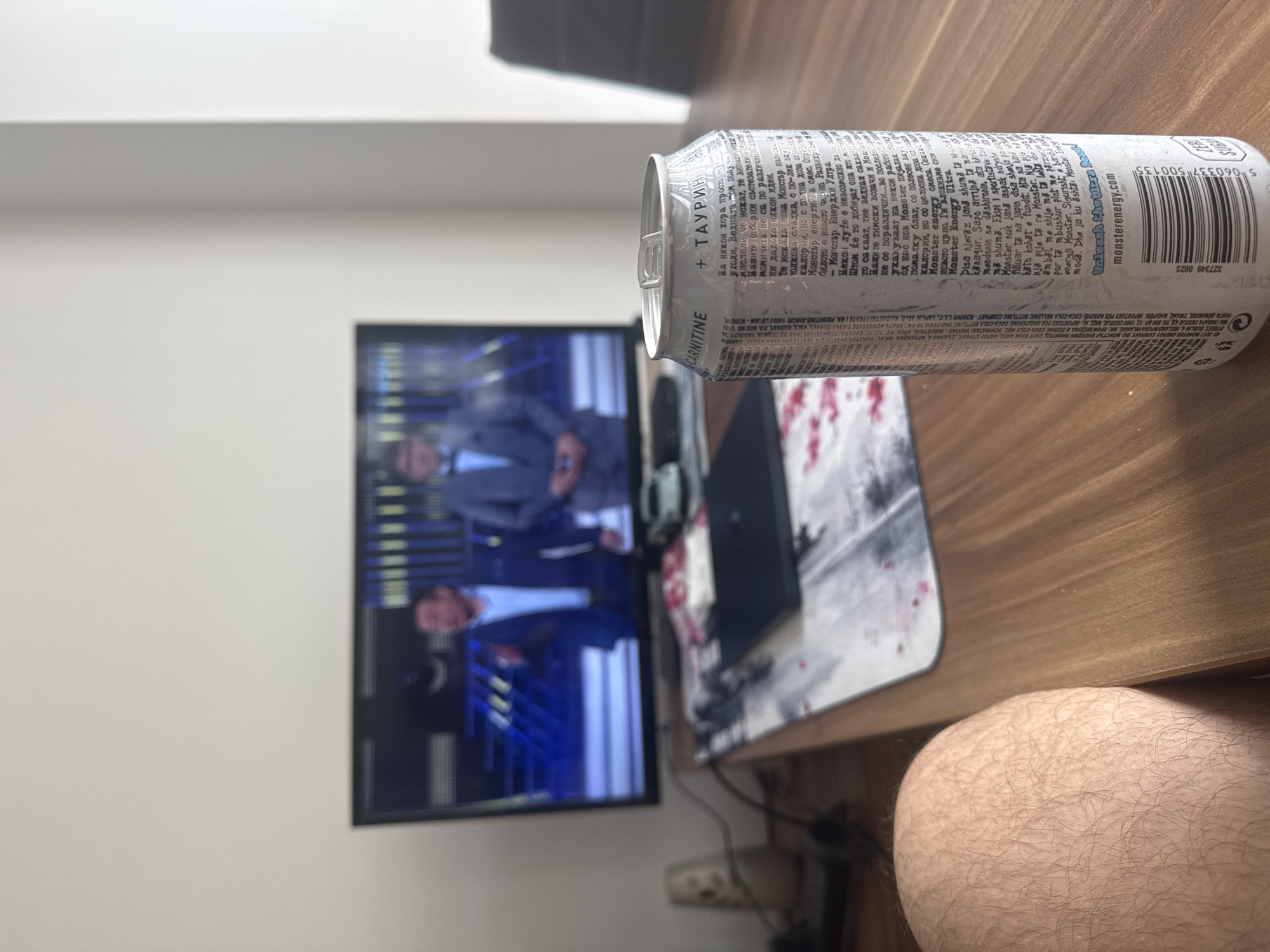The image presents a cluttered desk setup. A partially visible leg with hairy skin is in the bottom right. A silver Monster energy drink can is prominently positioned in the upper center, appearing to float mid-air. Behind it is a computer setup, including a black keyboard, mouse, and a mousepad with a mountain design. A television screen displaying two men in suits is in the background. The wall behind the TV is plain white. The desk surface appears to be made of light-colored wood. The location is assumed to be in Sofia, Bulgaria, based on the Cyrillic alphabet visible on the Monster can.