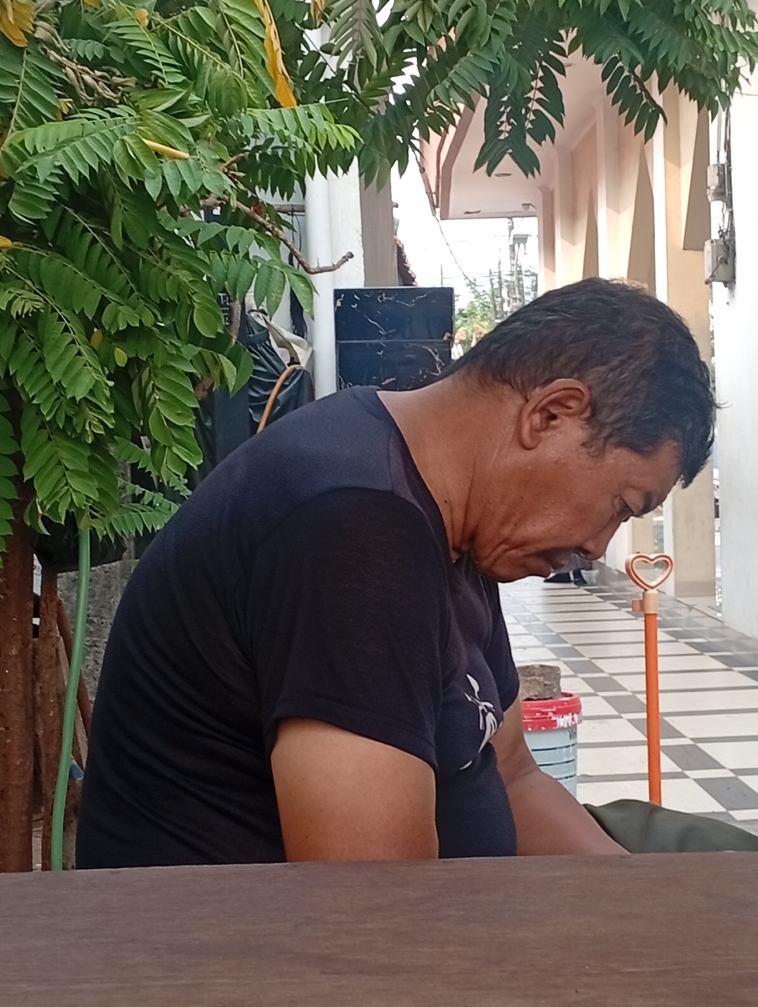 A man is sitting at a wooden table in what appears to be a courtyard or an outdoor waiting area. He is wearing a black t-shirt and his head is bowed as if he is resting or contemplating. The background shows a tiled floor with a checkered pattern, a walkway with decorative pillars, and lush green foliage from a tree. In the distance, there is a building with light-colored walls and a covered walkway. A small orange object with a heart shape at the top is visible to the right of the man. The image suggests a peaceful and somewhat secluded setting within a city environment in Bekasi, Indonesia, given the foliage and architecture.