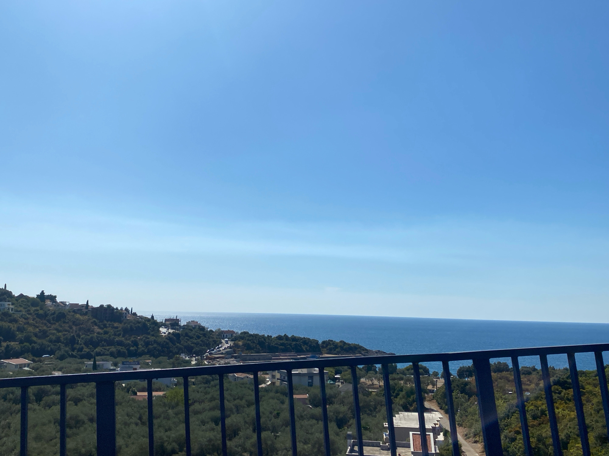 This is a landscape photograph taken from a high vantage point overlooking the Adriatic Sea in Kunje, Montenegro. The foreground is dominated by a dark blue metal railing, typical of a balcony or terrace. Through the railing, a dense, green olive grove is visible, with hints of buildings and roads interspersed within the foliage.
Beyond the trees and structures, the main subject is the vast expanse of the deep blue Adriatic Sea, stretching to the horizon. The coastline is visible on the left and in the mid-ground, characterized by a hilly terrain covered in lush vegetation and dotted with buildings, some appearing to be houses or villas. A road winds its way along the coast, with a few vehicles visible. The sea is calm, reflecting the clear, bright blue sky above. The sky itself is a gradient of pale blue at the horizon, becoming a deeper blue towards the top of the frame, with a faint haze visible near the sea. The overall impression is one of a serene coastal landscape on a sunny day.