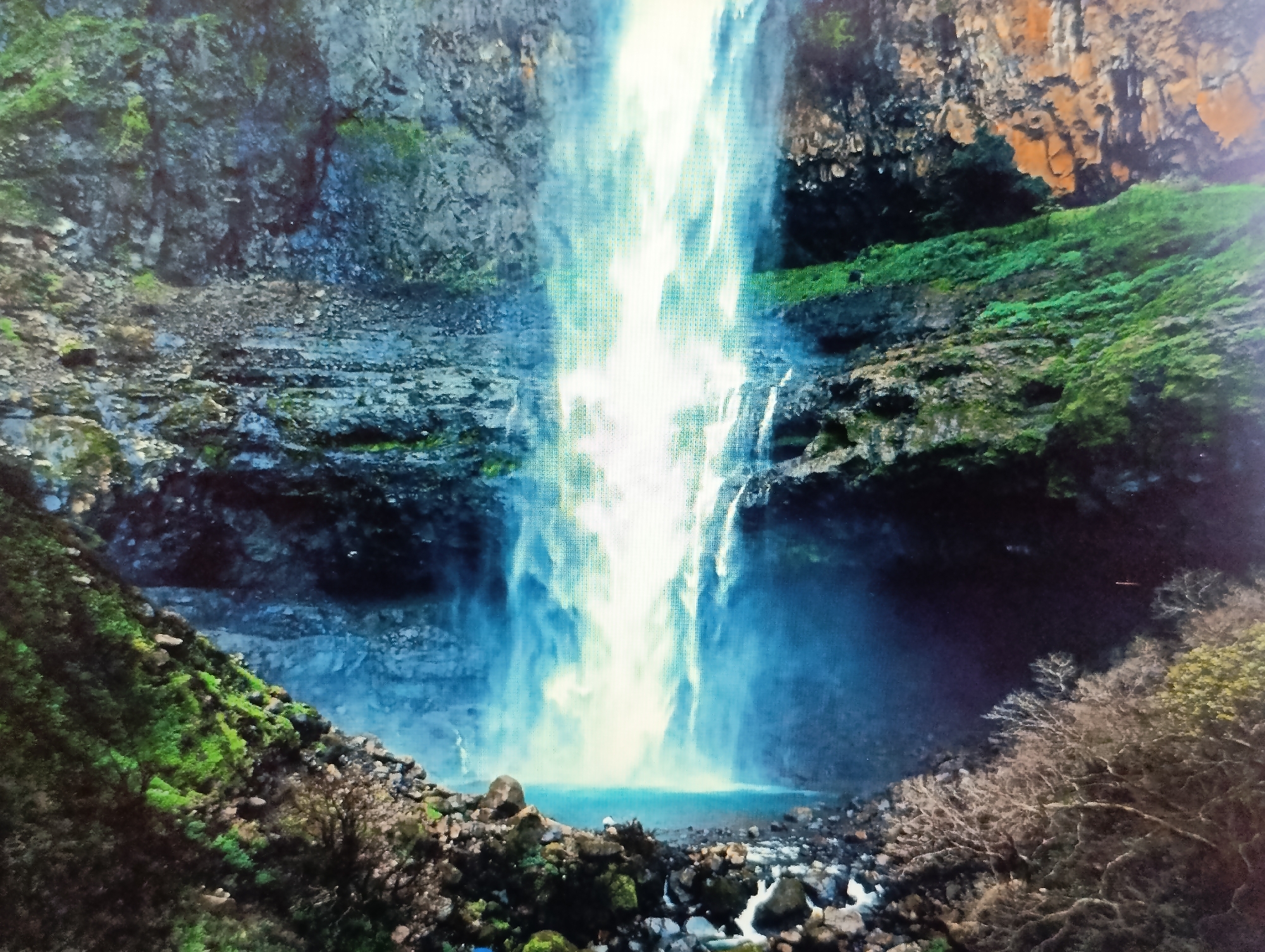 The image shows a powerful waterfall cascading down a rocky cliff face into a pool of water at its base. The surrounding environment is lush and green, with vegetation clinging to the rocks and slopes. The water appears bright and energetic, with a mist rising from the impact at the bottom. The scene is captured from a perspective that emphasizes the scale and force of the waterfall. There are no people visible in the image. The location is not in Dubăsari town council, Moldova, as this image depicts a natural landscape unrelated to that region.