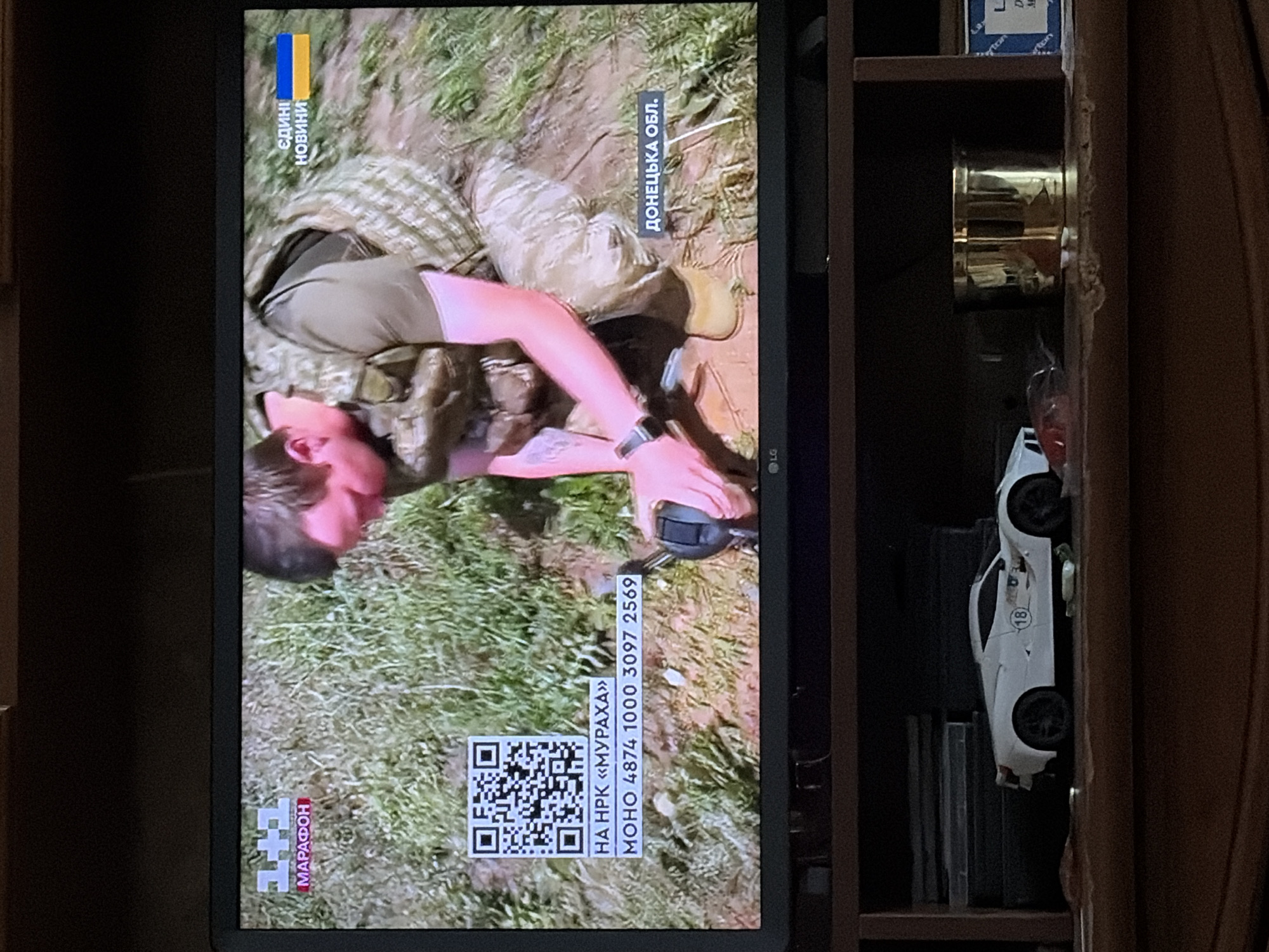 The image shows a television screen displaying a news broadcast. On the screen, a man in military fatigues is shown kneeling in mud. To the left of the man, there is a QR code and some text in Ukrainian that reads "НА НРК «МУРАХА» МОНО 4874 1000 3097 2569". To the right of the man, there is text in Ukrainian that reads "ДОНЕЦЬКА ОБЛ.". The television is placed on a wooden shelf, and in front of the television, there is a white toy car with the number 18 on it. Behind the television, there are some books or boxes. The overall environment is dimly lit.