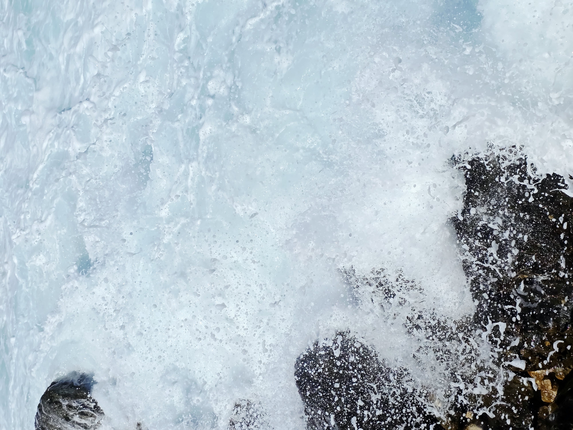 The image shows a close-up, high-angle view of a wave crashing against dark rocks on the coast of Chania, Greece. The water is a frothy, light blue and white, with numerous small bubbles and splashes visible. The dark rocks, wet from the impact of the waves, are textured and appear to be a natural formation along the coastline. The scene is dominated by the dynamic movement of the water, creating a sense of power and nature's force. There are no people or any man-made objects visible in the frame. The environment is exclusively oceanic and rocky.