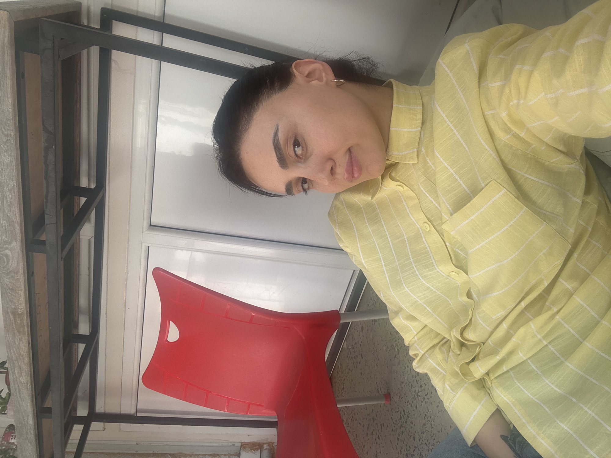 A portrait of a person is taken from a high angle. The individual has dark hair and is wearing a light yellow button-down shirt with a white grid pattern. A bright red plastic chair is visible in the foreground. The background includes a wall with vertical white panels and a black metal shelving unit. The location is Tbilisi, Georgia.