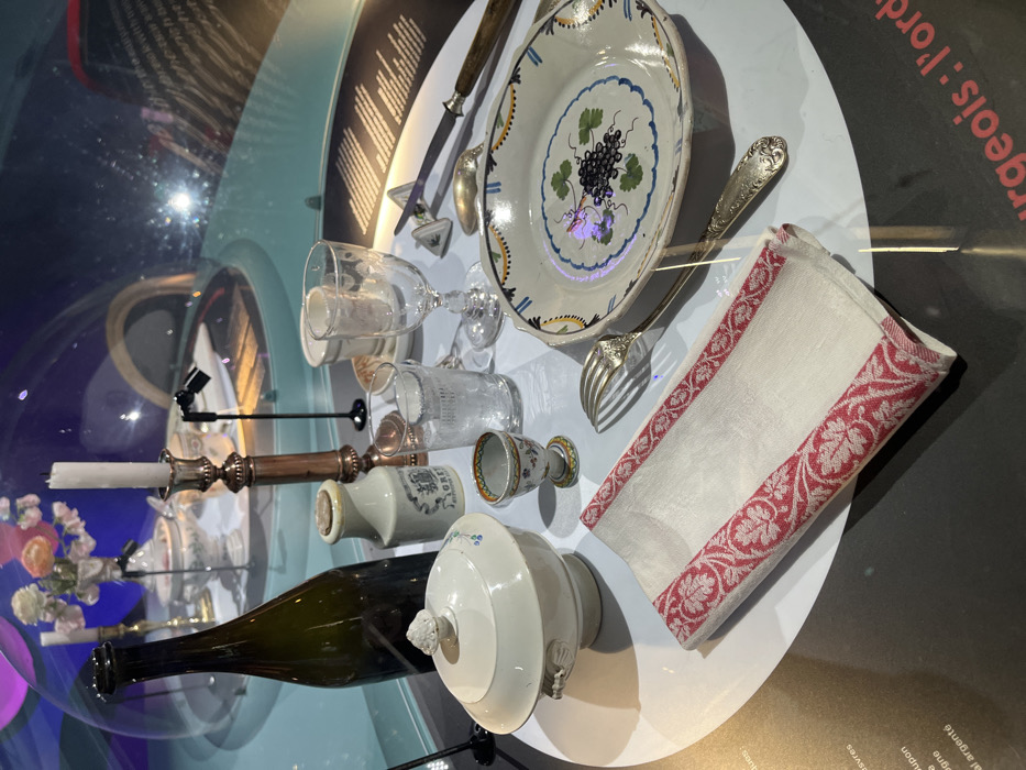 This image displays a historical dining table setting presented as an exhibit within the Cité de la Gastronomie et du Vin in Dijon, France. The objects are arranged on a circular white platform, partially enclosed by a clear, reflective acrylic dome.
In the foreground, a folded white linen napkin features a prominent red and white intricate pattern along its border. To its left, a white ceramic lidded soup tureen with decorative handles and faint blue floral motifs rests. Behind the tureen, a dark green glass bottle stands upright. A small, multi-colored ceramic egg cup with floral designs is positioned between the napkin and the tureen.
Further back in the display, a plain white stoneware jar, partially labeled "GREY," is placed near a tall brass candlestick holding a lit white candle. To the right, a clear glass tumbler and a stemmed wine glass are arranged. An oval-shaped ceramic plate dominates the right side, showcasing a hand-painted central motif of dark grapes and green leaves, encircled by blue and yellow decorative bands. A silver fork and a knife with a wooden handle are positioned next to the plate, accompanied by an additional silver spoon and a small white triangular condiment holder.
The background features abstract blue and purple ambient lighting, creating reflections on the dome. Another, smaller table setting is discernible further back within the enclosed space. Partially visible French text on the dark perimeter surrounding the white display platform indicates informational labeling. The scene is devoid of people.
