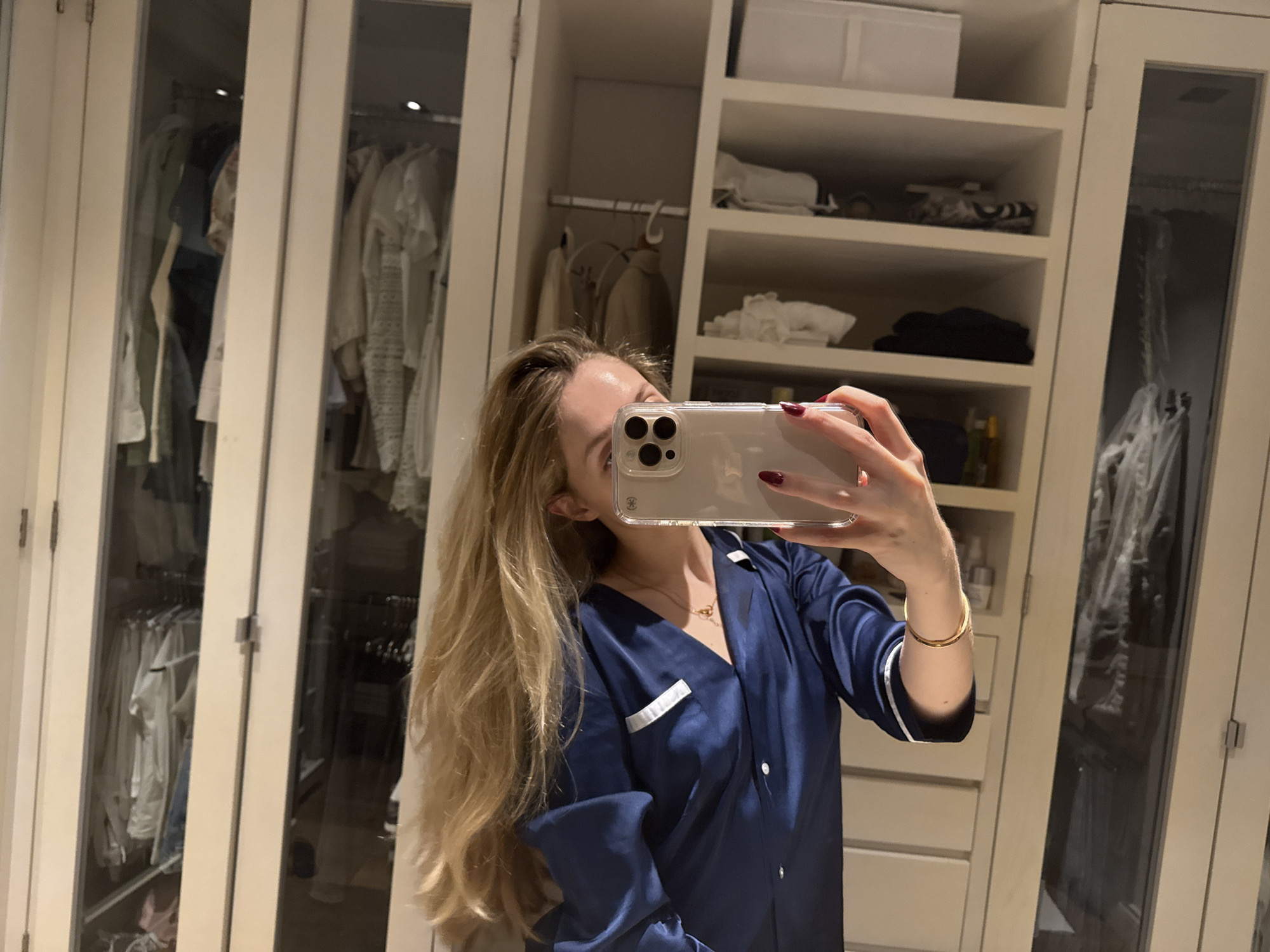 A woman is taking a selfie in front of a closet. The closet appears to be built-in, with white doors and shelves. Inside the closet, there are clothes hanging on hangers and items stacked on shelves. The woman is wearing a dark blue pajama set with white piping. She has long blonde hair and is wearing a gold necklace and bracelet. The lighting is soft and warm. The location is most likely a home in Mexico City, Mexico.