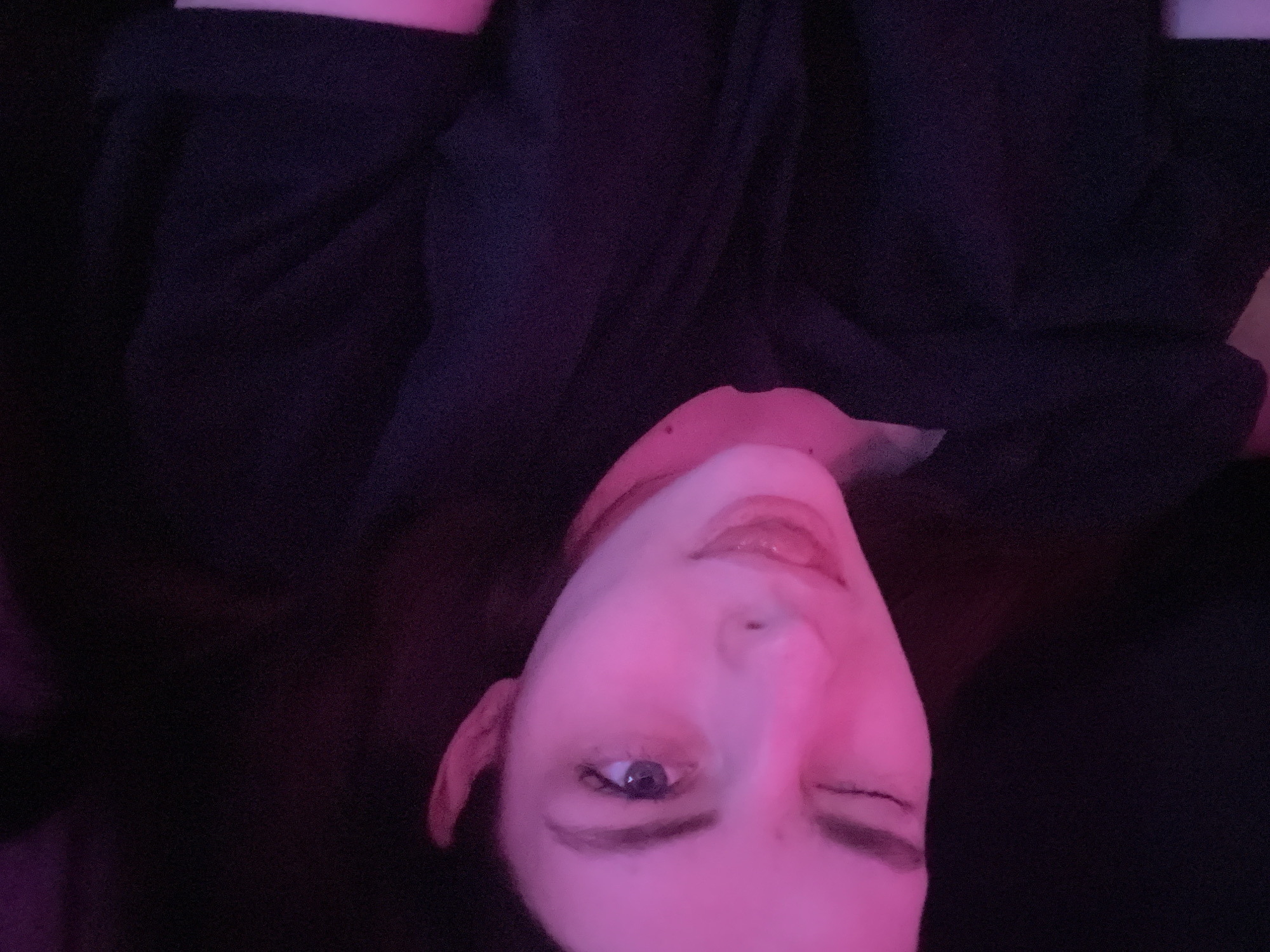 A selfie of a woman with one eye open and one eye squinted is illuminated by a pink light. She is wearing a dark-colored garment. The location is unknown.