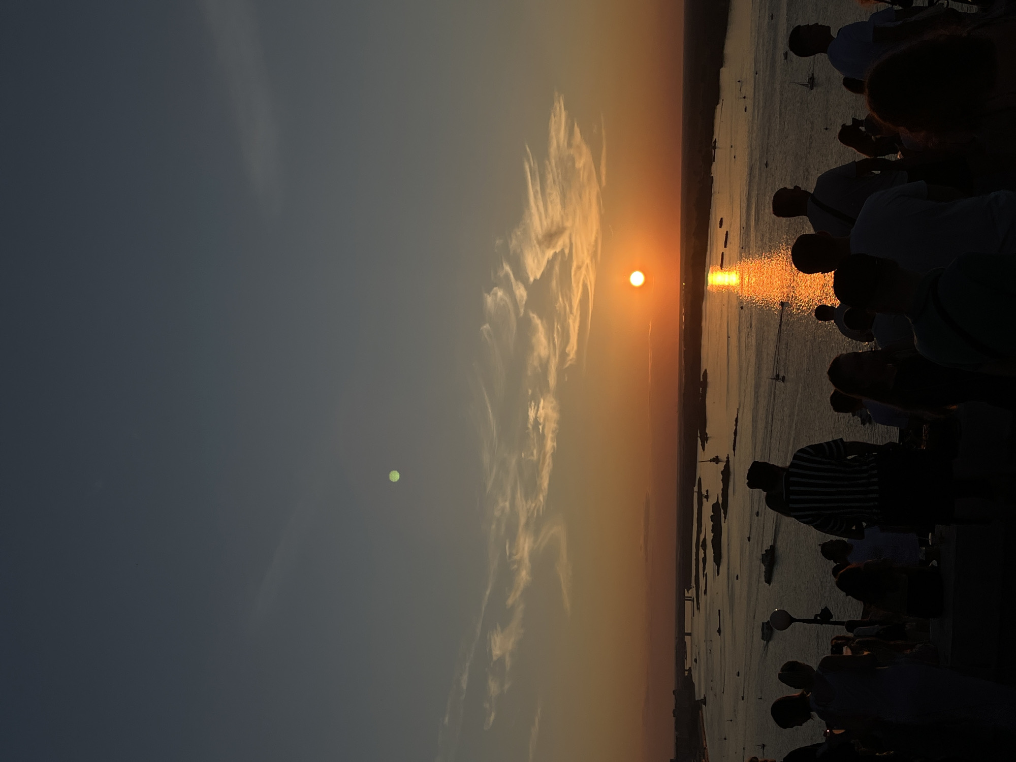 The image captures a sunset over the Volga River in Nizhny Novgorod, Russia, viewed from a high vantage point. In the foreground, silhouetted against the warm glow of the setting sun, is a crowd of people gathered to watch the spectacle. They are standing on an elevated area, likely a viewpoint or observation deck, overlooking the river.
The river itself is wide and appears to be busy with various boats, including larger vessels and sailboats, dotting its surface. The setting sun casts a brilliant, shimmering reflection on the water, creating a golden pathway. In the distance, the cityscape of Nizhny Novgorod is visible, with bridges spanning the river and structures hinting at its urban presence. The sky is a gradient of deep blue at the top, transitioning to warm oranges and yellows near the horizon, with wispy clouds adding texture to the scene. A faint, greenish orb is visible in the upper part of the sky, possibly a celestial body or an artifact of the camera. The overall atmosphere is one of serene observation and appreciation of the natural beauty of the sunset.