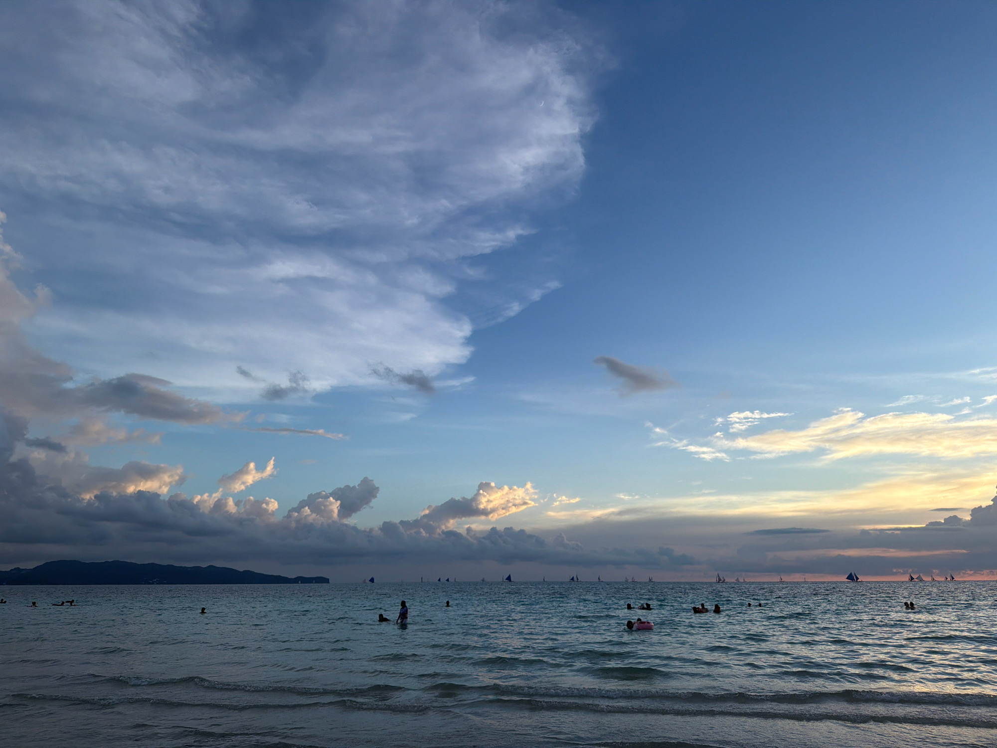 The image depicts a wide, panoramic view of Boracay Island's beachfront in Malay, Philippines, during a sunset. The sky is a gradient of blue, transitioning to hues of orange and yellow near the horizon, with a dramatic arrangement of clouds. Several sailboats are visible dotting the ocean, and numerous people are in the water, some swimming and others relaxing. In the foreground, gentle waves lap against the sandy shore. The overall impression is of a serene and picturesque coastal scene at dusk.