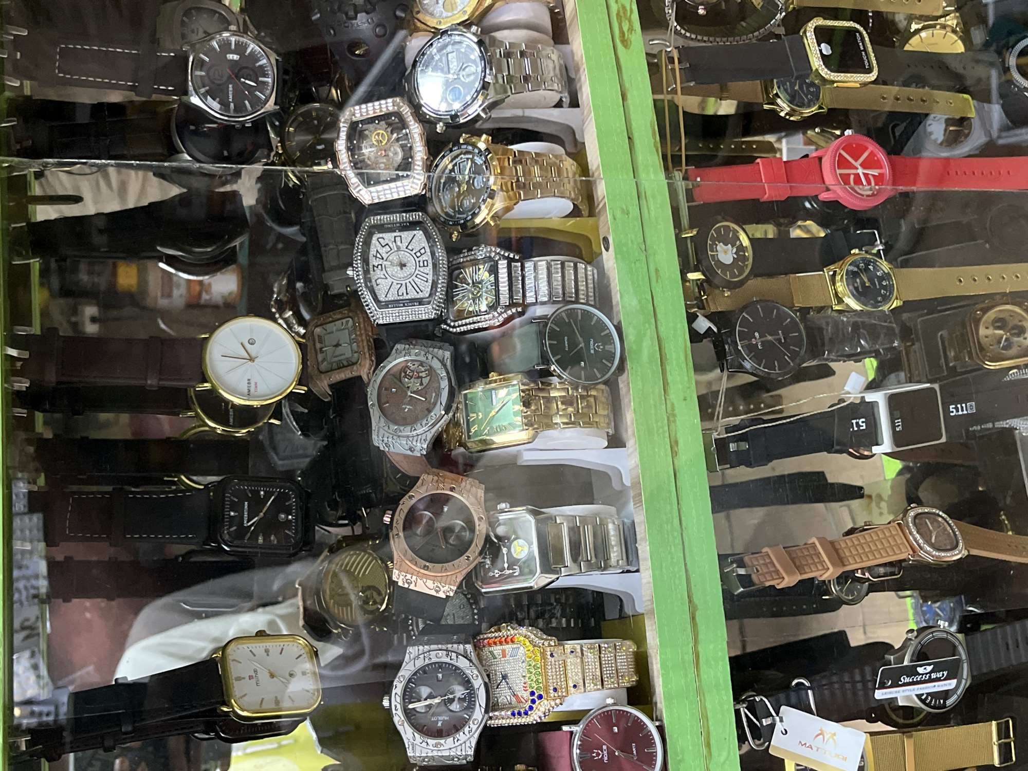 In Bukuru, Nigeria, a stall displays a variety of watches behind a glass enclosure. The watches are arranged on multiple shelves and hanging displays. Prominently featured are several gold-colored watches with white or patterned faces, some embellished with rhinestones. There are also watches with black bands and faces, as well as stainless steel models. Other visible watches include a red-strapped watch with a unique dial, a watch with a green face, and several watches with rectangular faces. Some watches have visible brand names like "MIHOO," "AMEBR," "YACHTING," and "TOUCH WATCH." The lighting suggests an indoor setting, likely a market or shop.