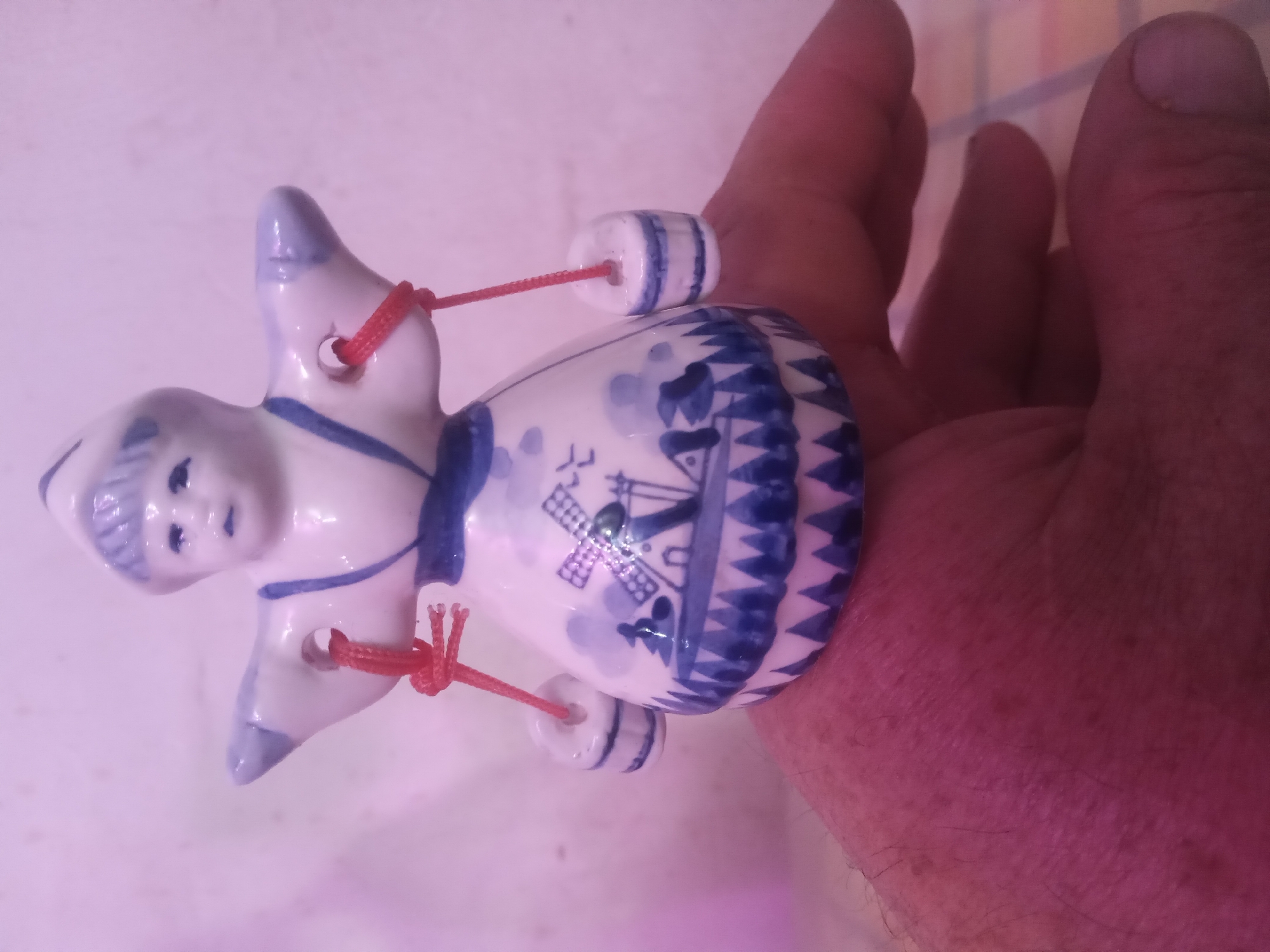 A white and blue ceramic figurine, designed in a style reminiscent of traditional Dutch pottery, is positioned centrally, resting on the upturned palm of an adult male's hand. The figurine depicts a figure, likely female, wearing a peaked cap and a dress adorned with blue patterns including a windmill, houses, trees, and a sawtooth border. Two small, cylindrical white and blue ceramic buckets are suspended by red strings from holes located in the figurine's outstretched, wing-like arms. The hand is open, with fingers slightly curled, and exhibits visible hair on the wrist and back. The skin tone of the hand appears pinkish due to the ambient magenta-hued lighting. The background is a plain, light-colored wall, also illuminated with a pinkish glow. Below the supporting hand, a subtle geometric pattern, possibly from a mat or tablecloth, is partially visible. Location: Unknown, Unknown.