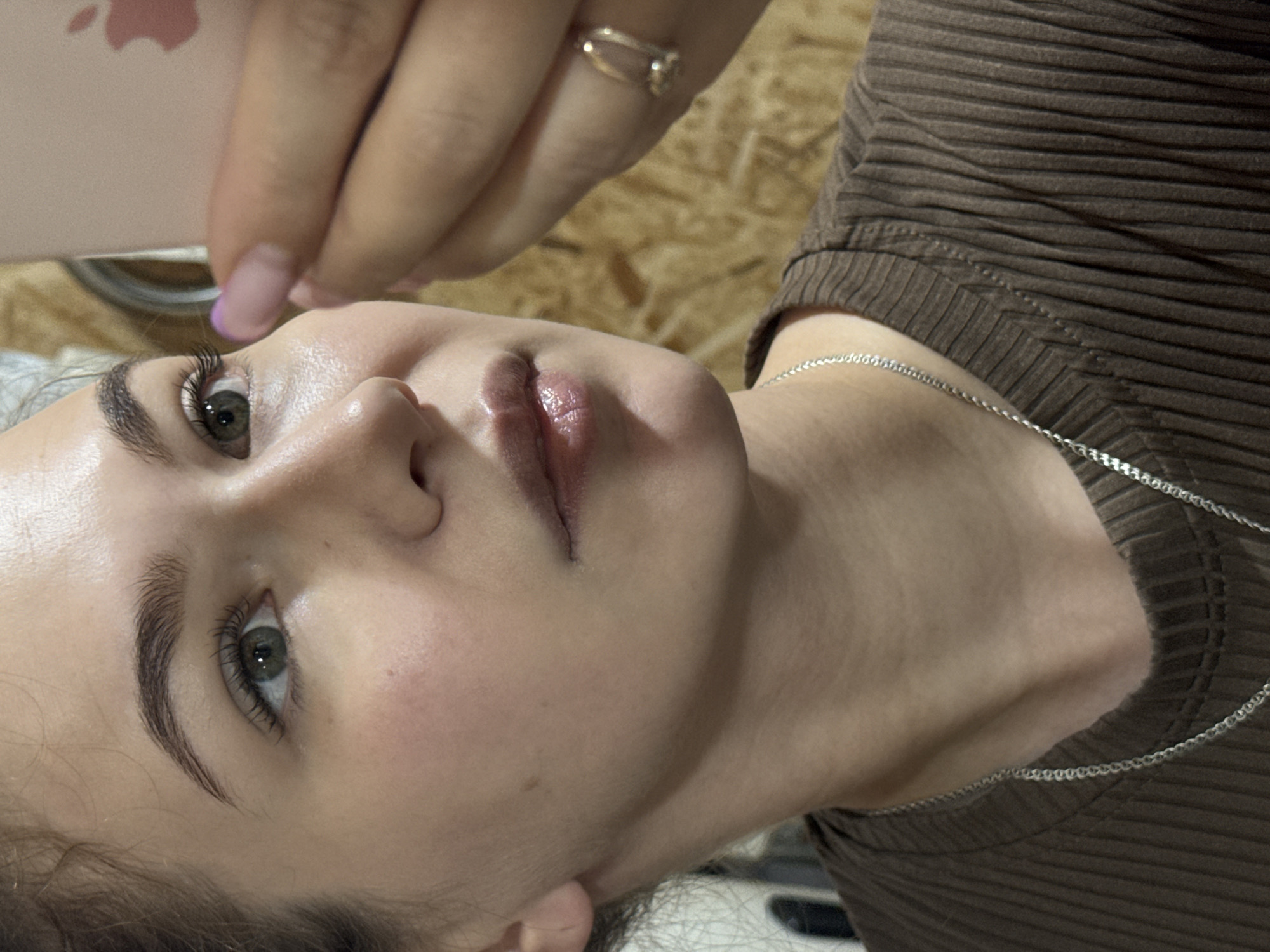 A close-up, angled shot depicts a woman lying on her back. Her face is pale with prominent eyelashes and arched eyebrows. Her lips are glossy. A hand wearing a gold ring is positioned near her forehead. She is wearing a brown, ribbed top and a silver chain necklace. The background is out of focus and reveals a textured surface that could be wood. The location is not discernible from the image.