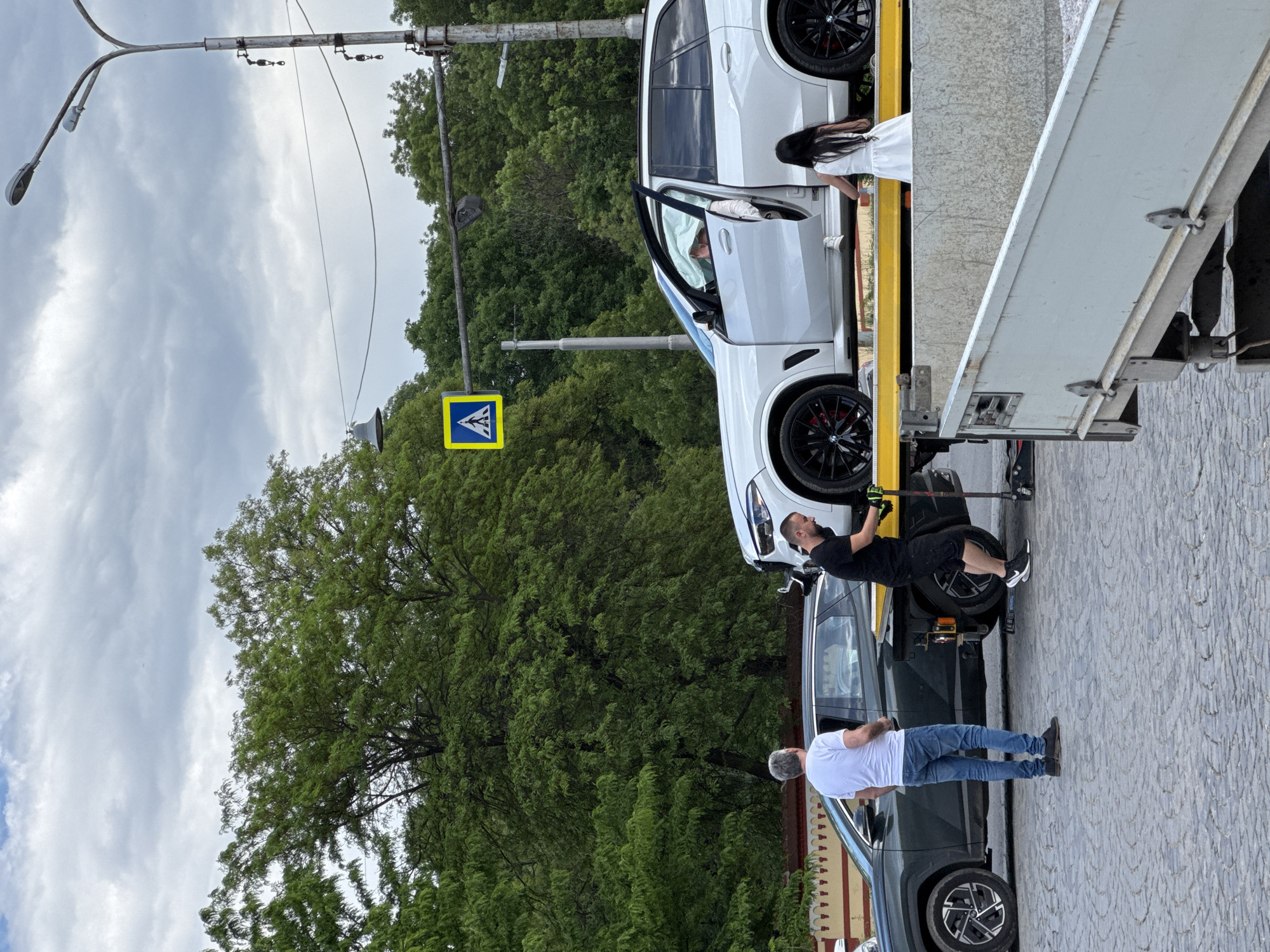 A white SUV is being towed in Sofia, Bulgaria. The car is on a flatbed tow truck on a cobblestone street, next to a pedestrian crossing sign. A man in a white shirt and blue jeans is standing near a grey car, watching the operation. Another man in a black shirt and shorts is working on the SUV. A woman in a white dress is standing on the tow truck bed, near the towed car. The background includes trees, a lamppost, and a cloudy sky.