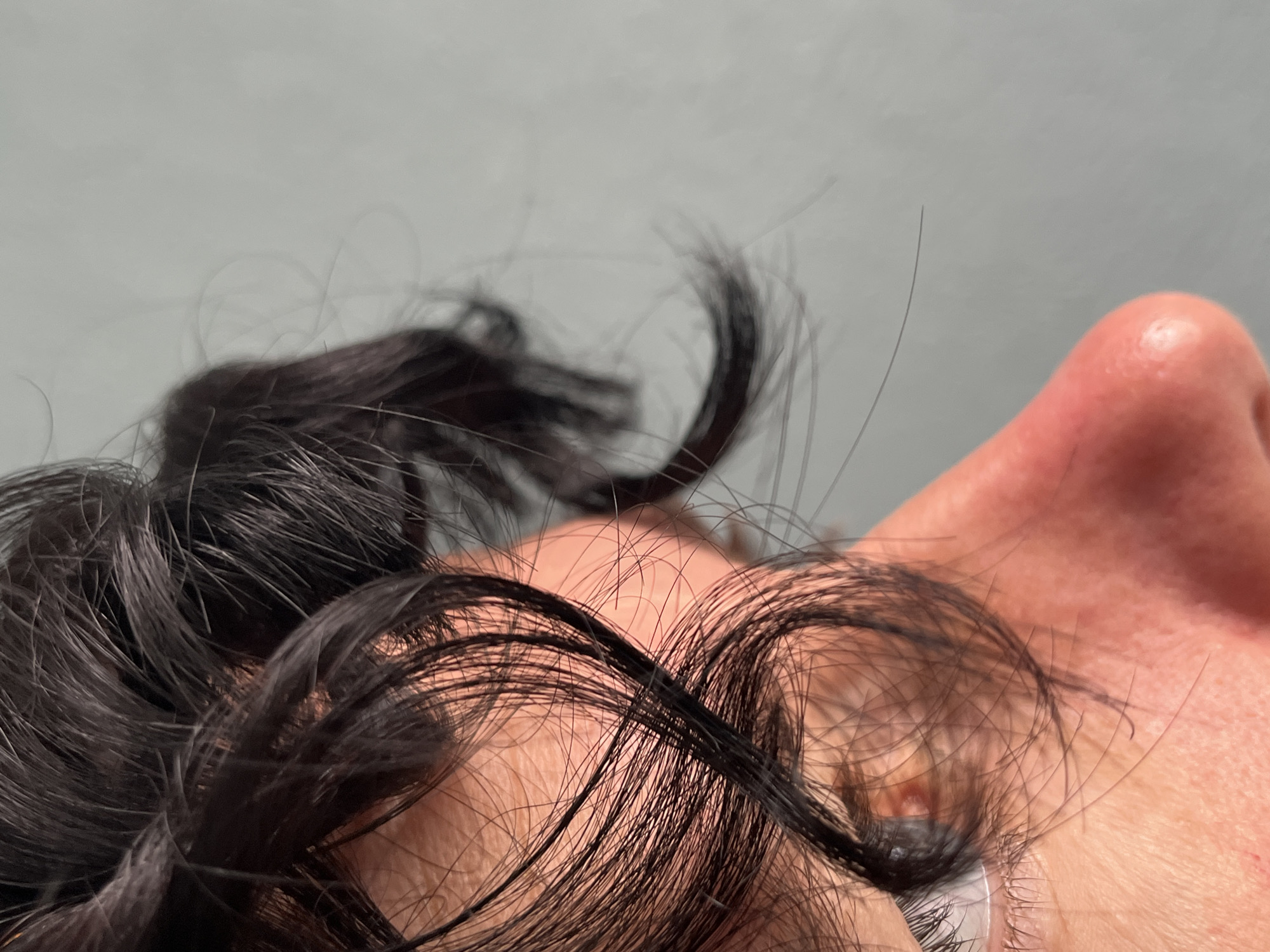 The image shows a close-up, profile view of a person's face, focusing on the eye and nose area. Dark, curly hair frames the face, with several strands falling across the eye. The skin is visible, with some texture and slight redness. The background is a plain, light grey wall. The location in ღრმაღელის თემი, Georgia, is not discernible from the image itself.