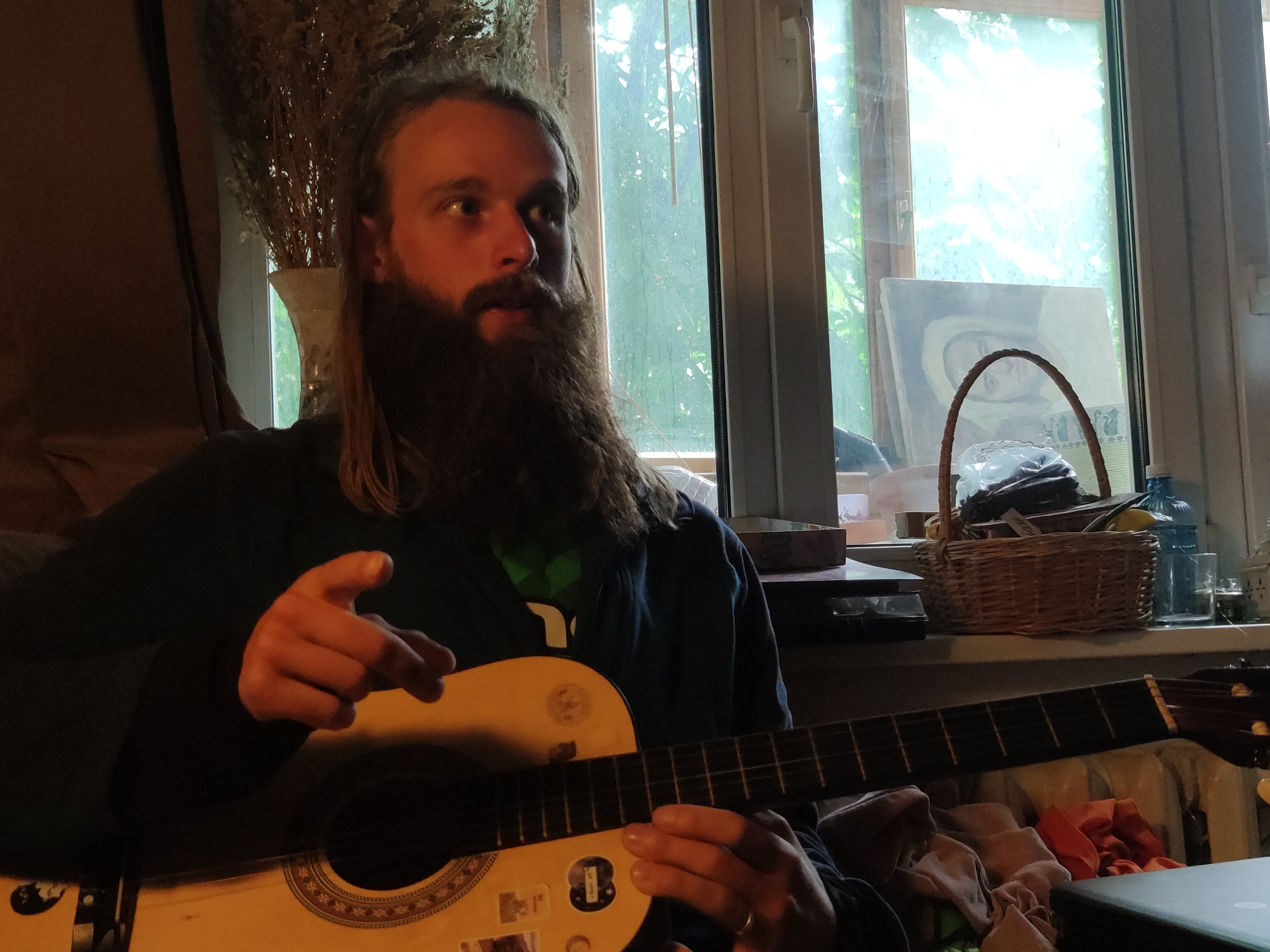 A bearded man with long hair is seated, playing an acoustic guitar. He is gesturing with his left hand. The background features a window with a cluttered windowsill. A basket, water bottle, and other items are visible on the sill. Behind the window, foliage suggests a green outdoor space. A vase with dried flowers is visible to the left. The location in the city of Moscow, Russia cannot be determined based on the image.